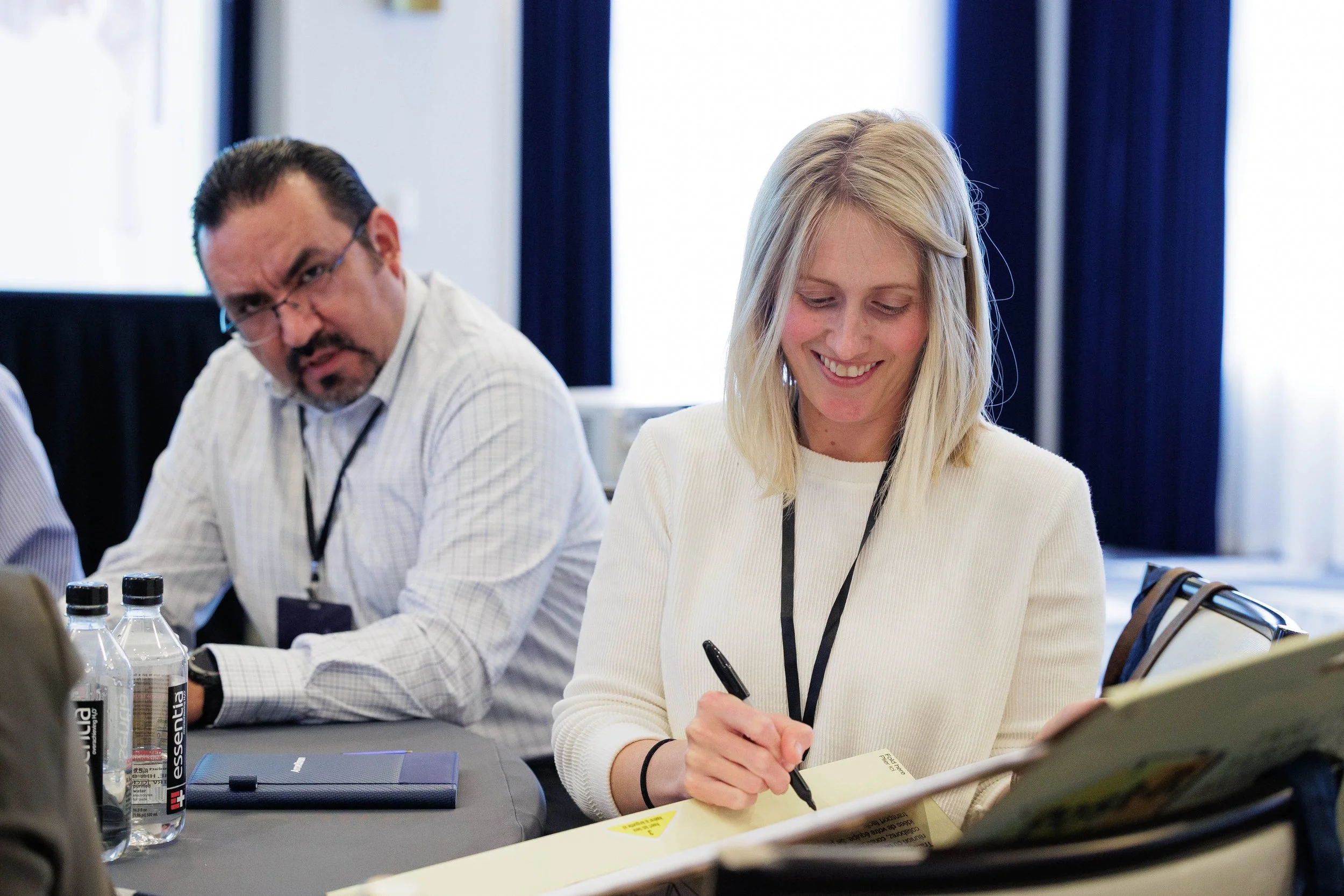 Woman smiling while writing notes at a Chicago conference seminar table as fellow attendee listens to presentation