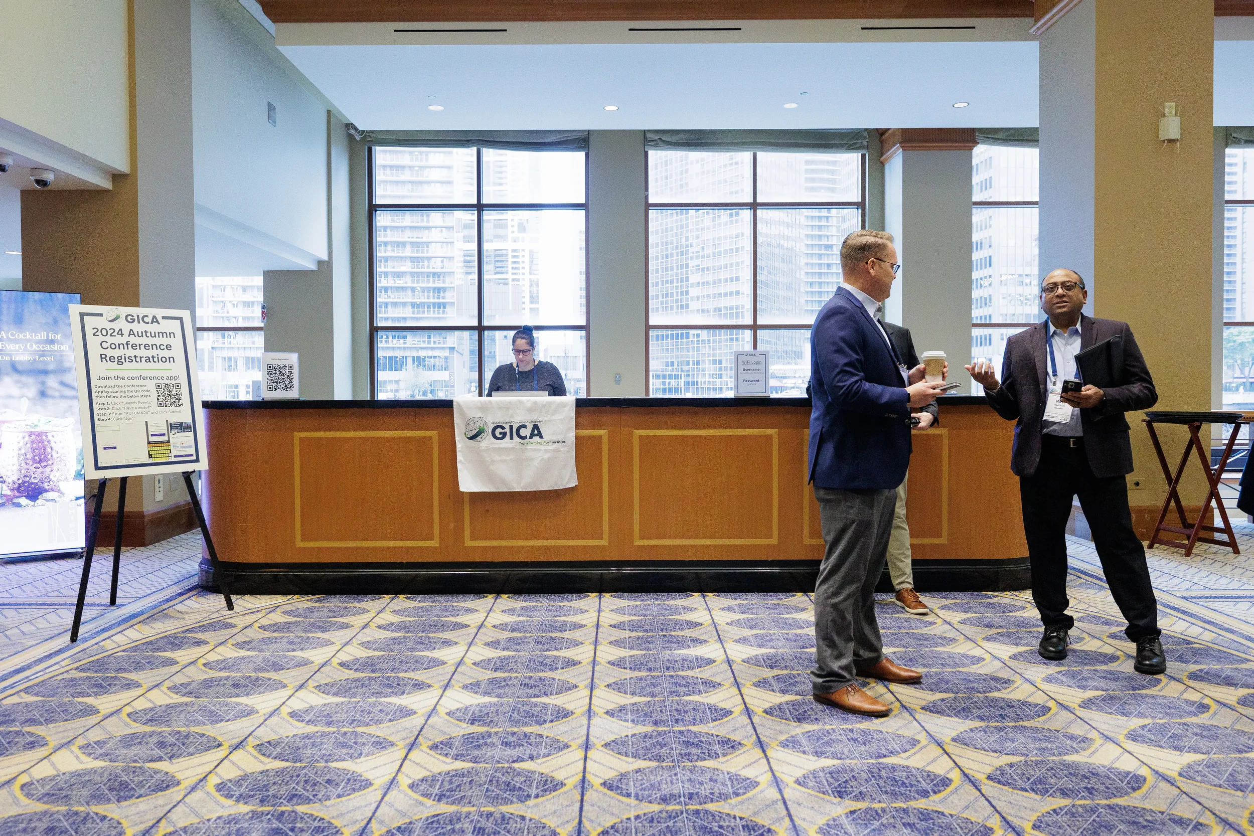 Two attendees chat near registration desk with Chicago skyline visible through windows at corporate conference