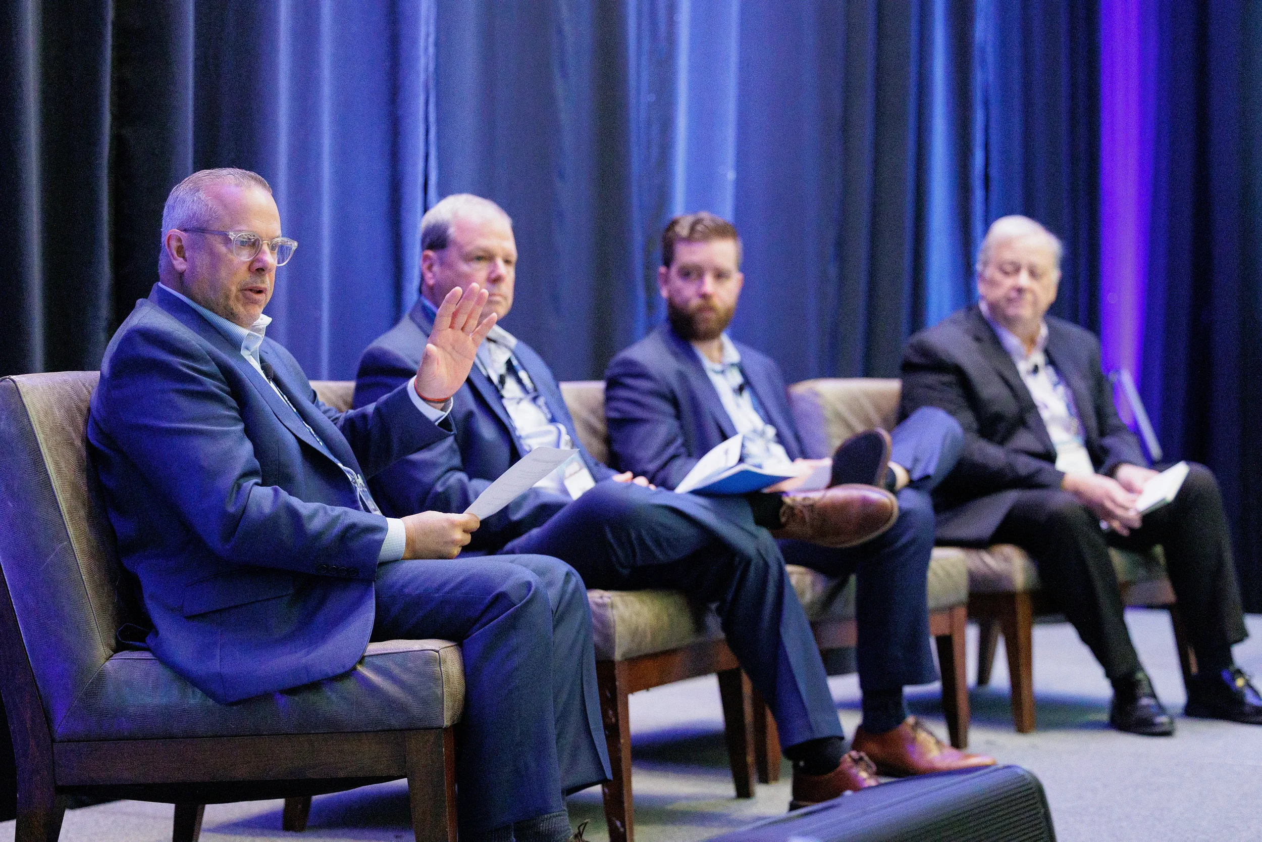 Four men in business suits sit in chairs on a stage with blue draped curtains during in a panel discussion in Orlando.