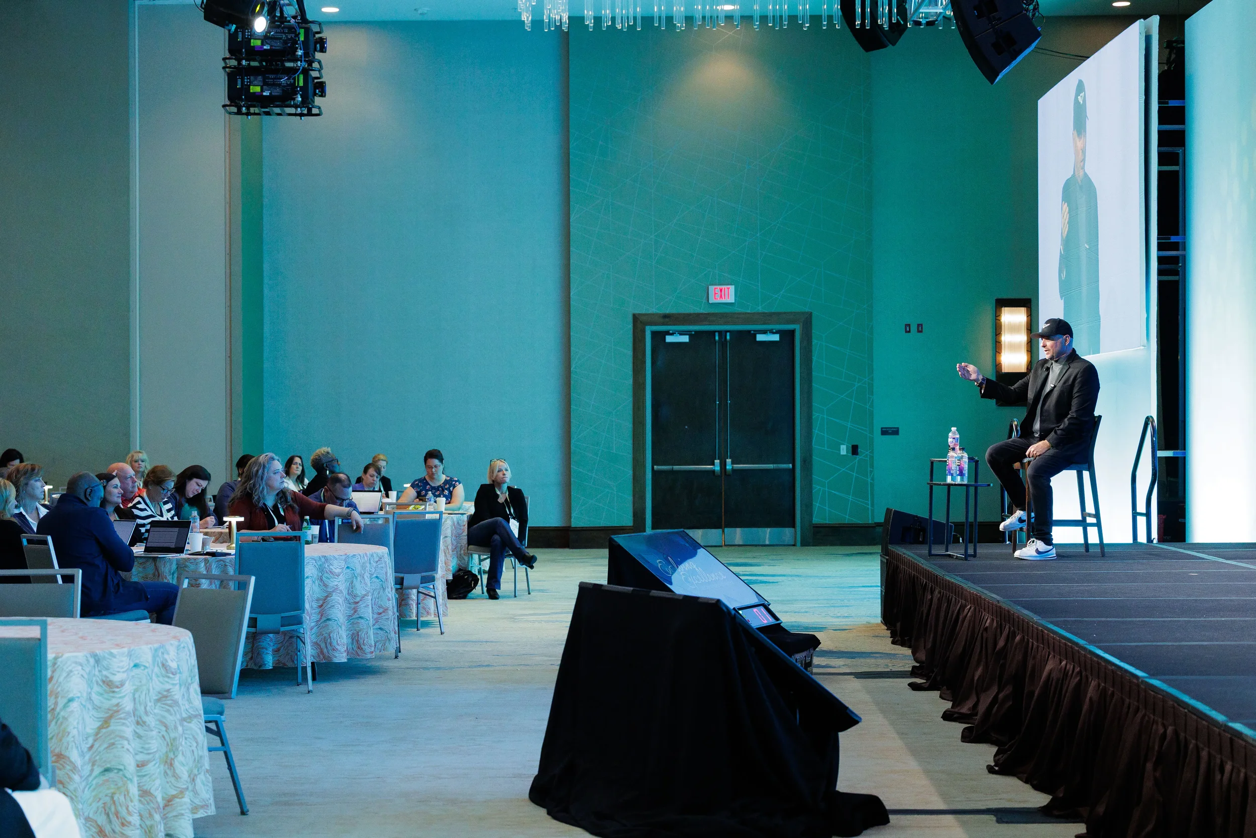 Speaker seated on stage addresses attendees at tables in teal-lit ballroom at Orlando corporate event
