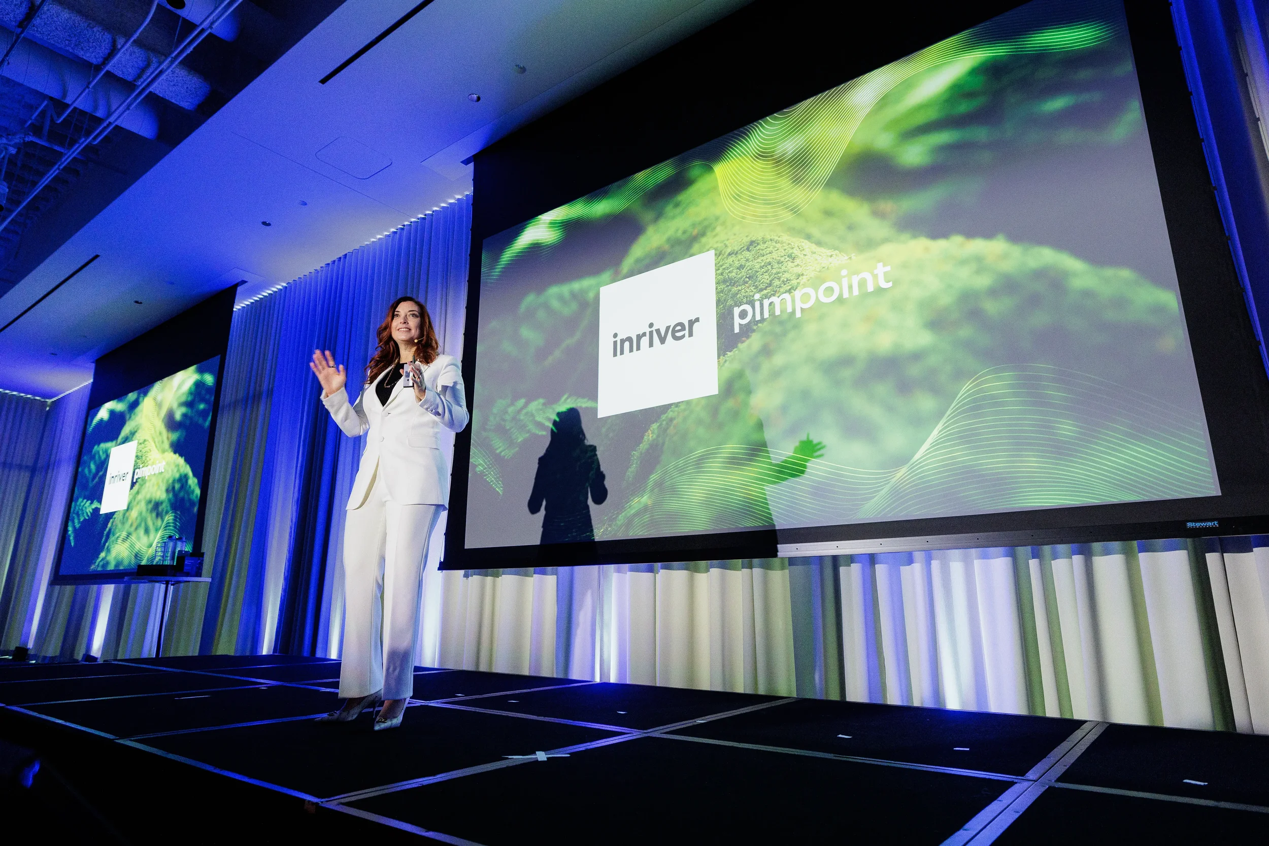 Speaker in white suit waves on stage beside green projection screen at Orlando corporate conference