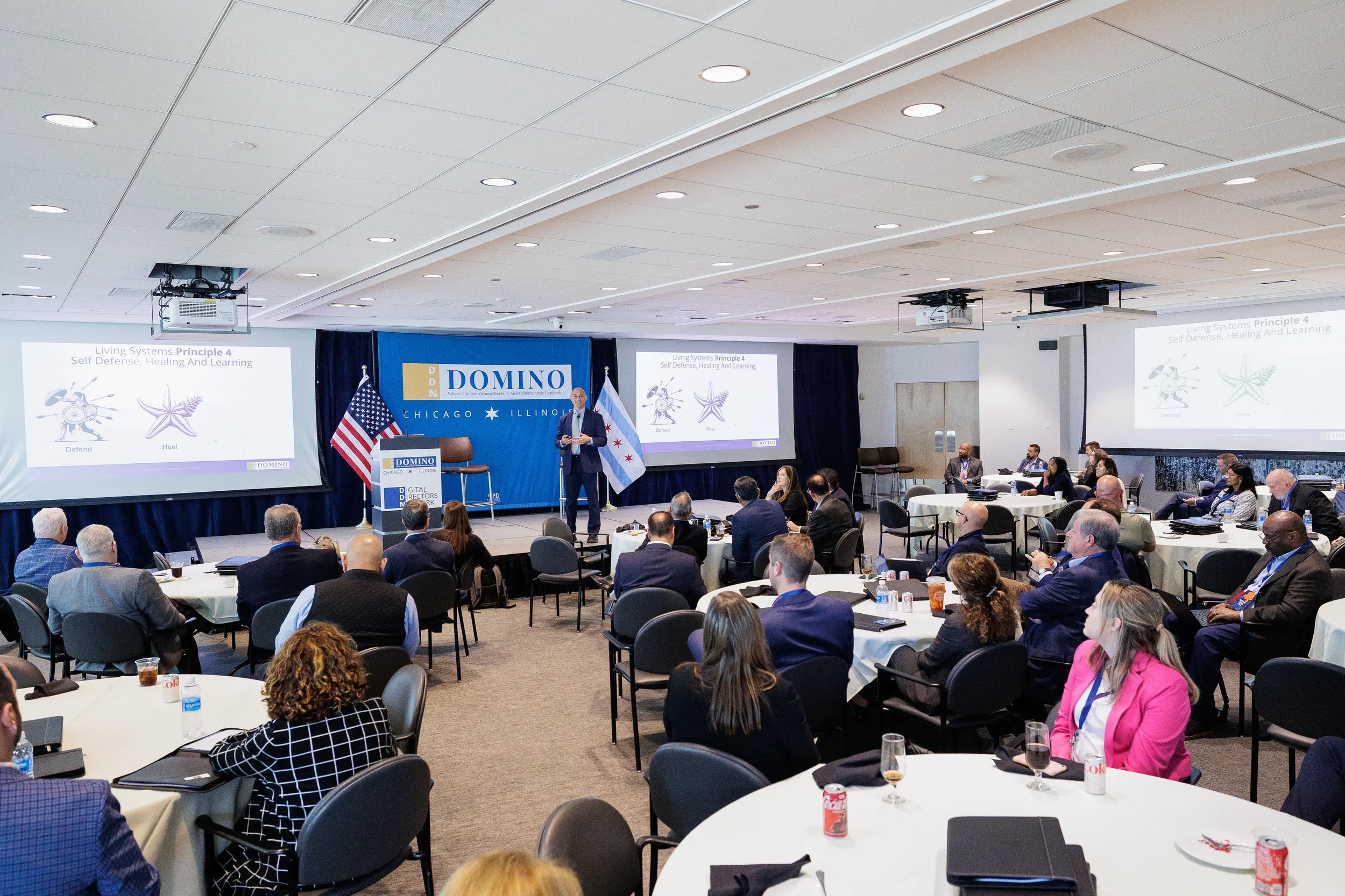 Speaker presents on branded stage to full audience at round tables with triple projection screens at Chicago conference