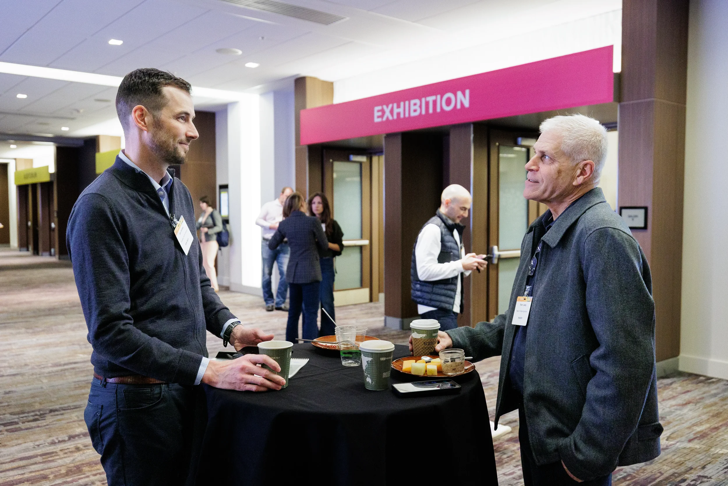 Two badged attendees chat over coffee at high-top table near exhibition entrance at Orlando corporate event