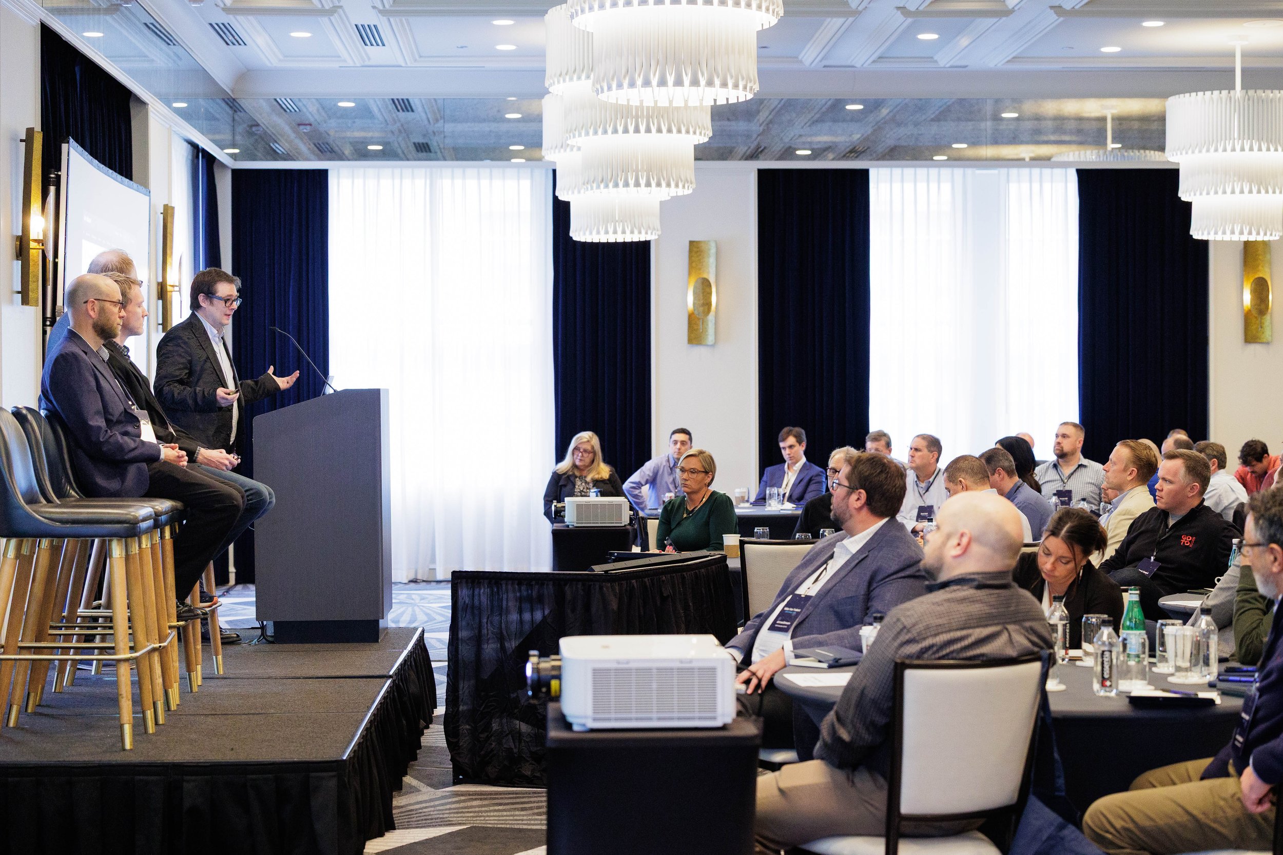 Speaker gesturing at podium while co-panelists and attentive audience members listen during a conference in Orlando
