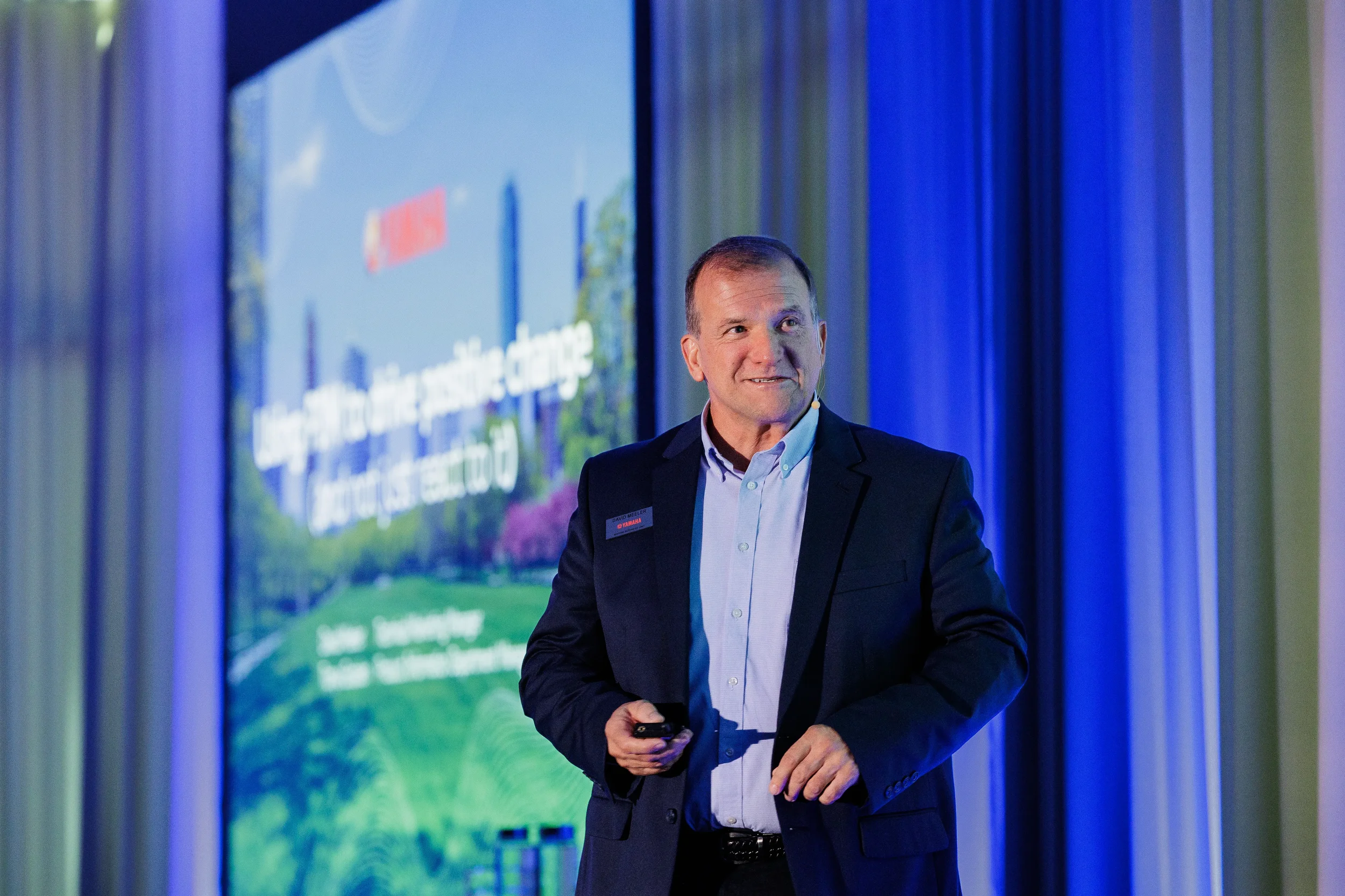 Smiling speaker with headset microphone holds clicker on stage before projection screen at Chicago conference