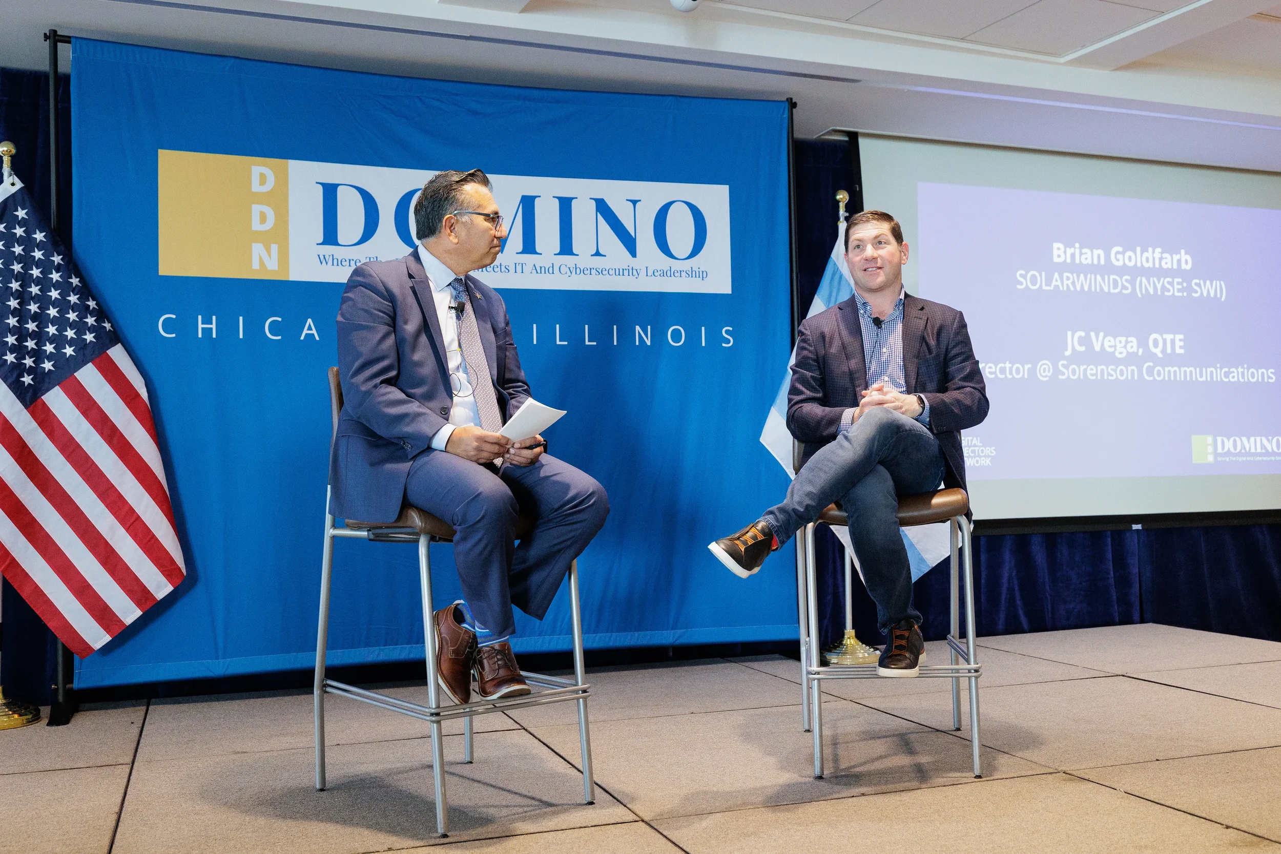 Two speakers seated for fireside chat on branded stage with flags at Orlando industry conference