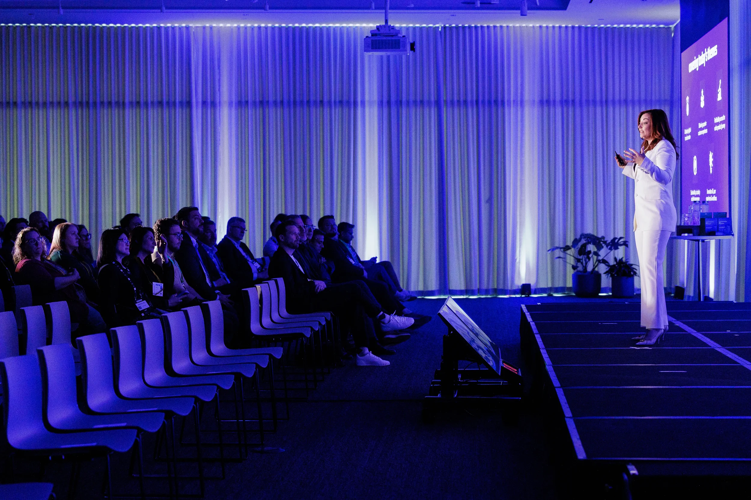 Speaker in white suit gestures to seated audience in blue-lit room at Orlando industry conference