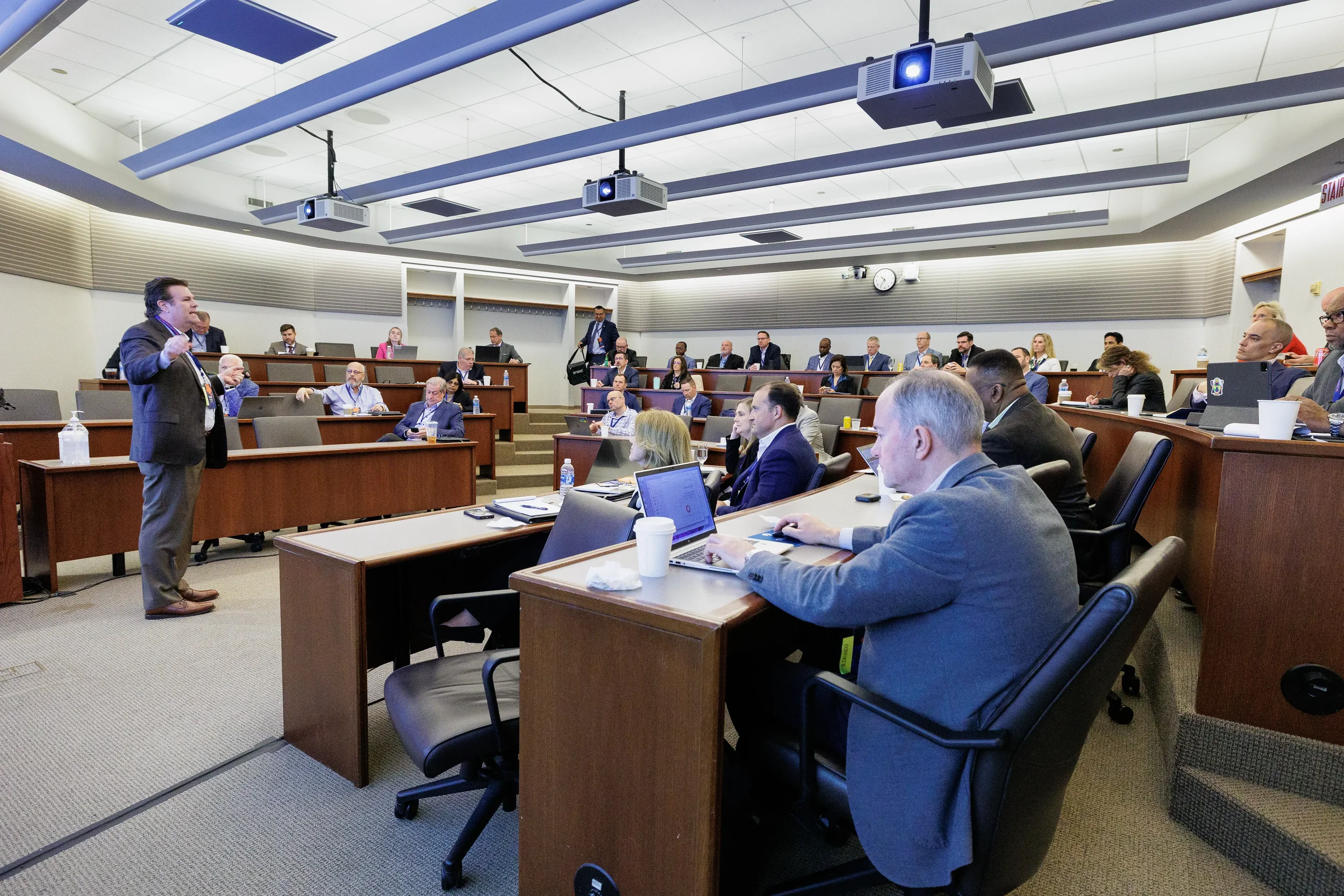 Male speaker gestures to full tiered classroom of attendees with laptops at Chicago corporate conference