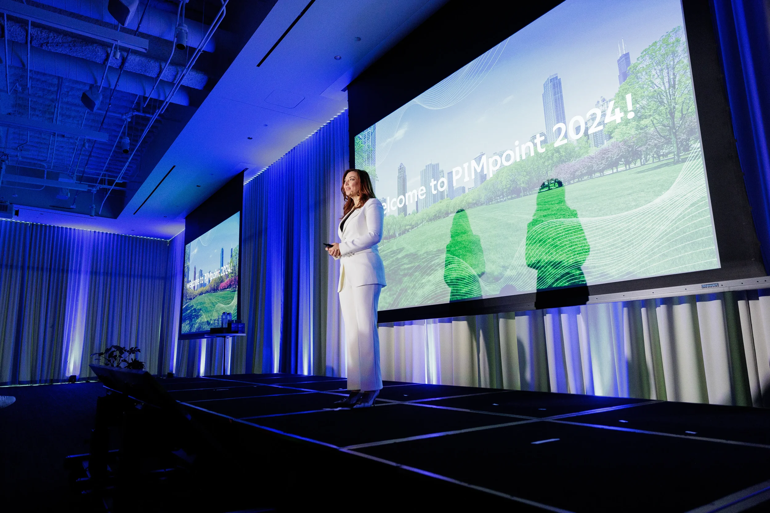 Speaker presents on stage with blue curtain lighting and large projection screens at Chicago industry conference