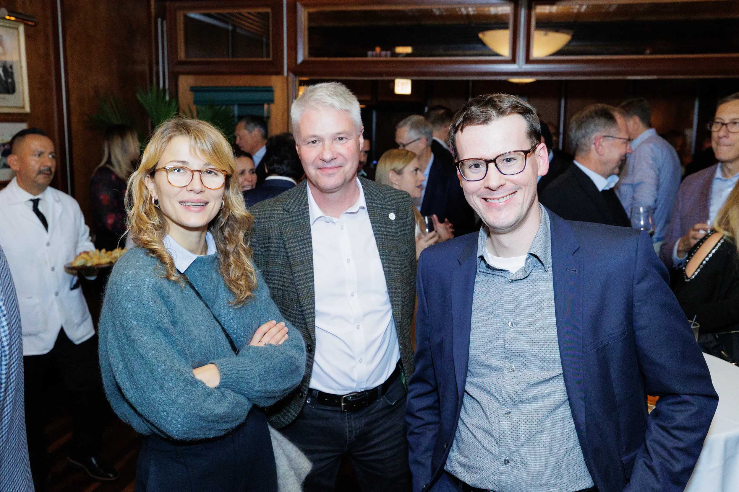 Three professionally dressed adults smile for a picture at an indoor networking event in Chicago with a crowded background.