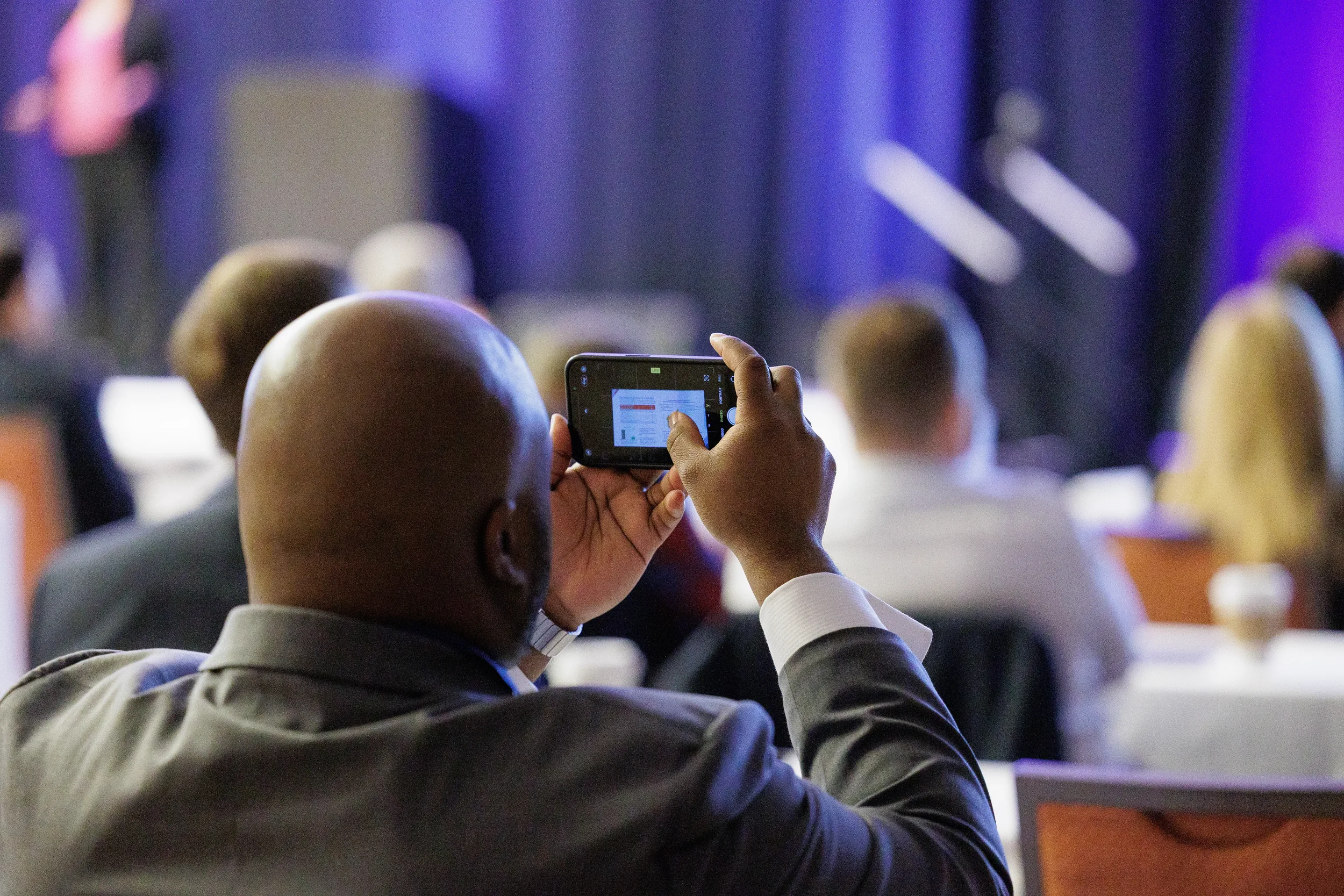A bald African American man in a suit takes a picture of a screen with a smartphone during a conference in Orlando.