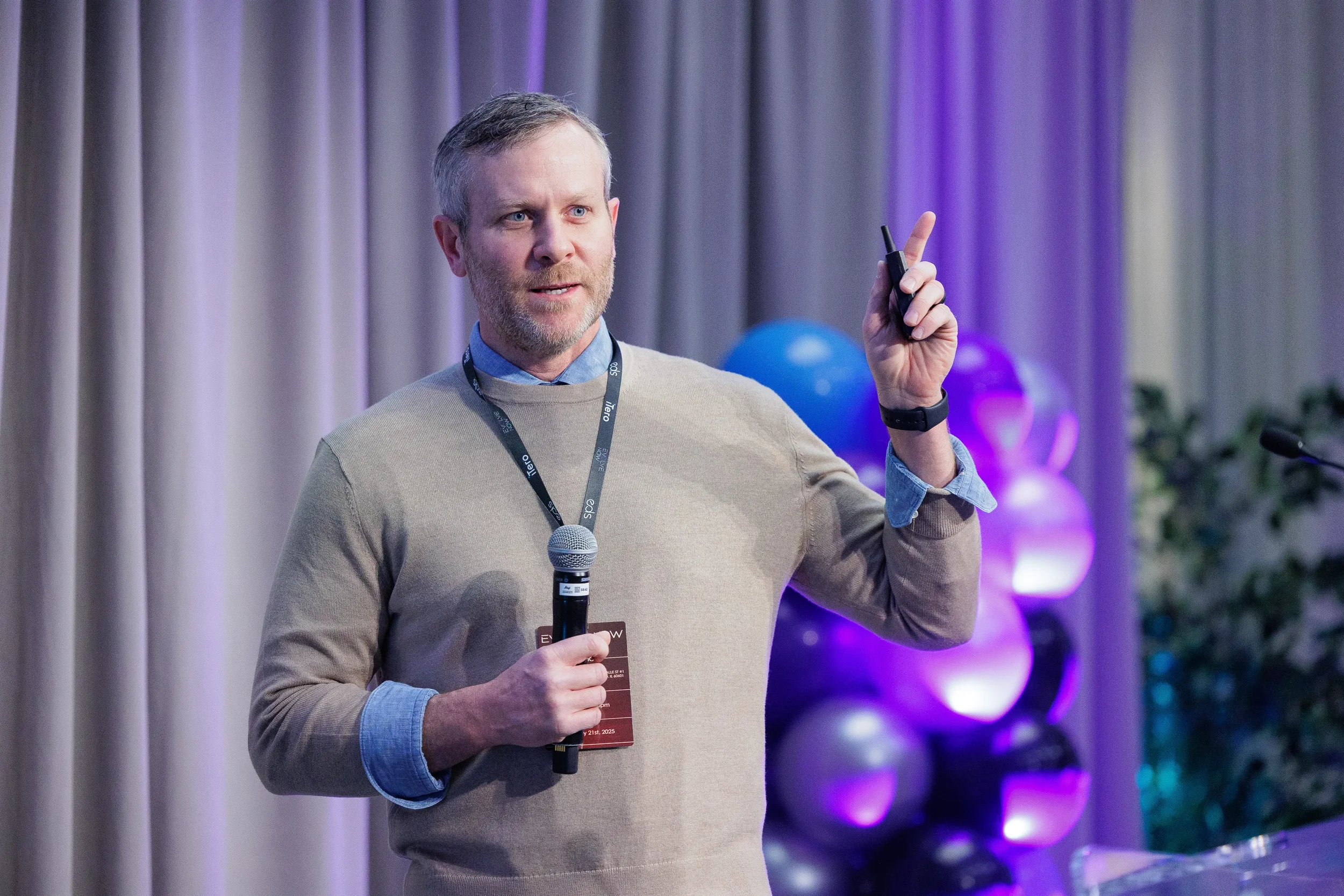 Lanyard-wearing speaker raises hand while presenting with balloon backdrop at Orlando industry conference