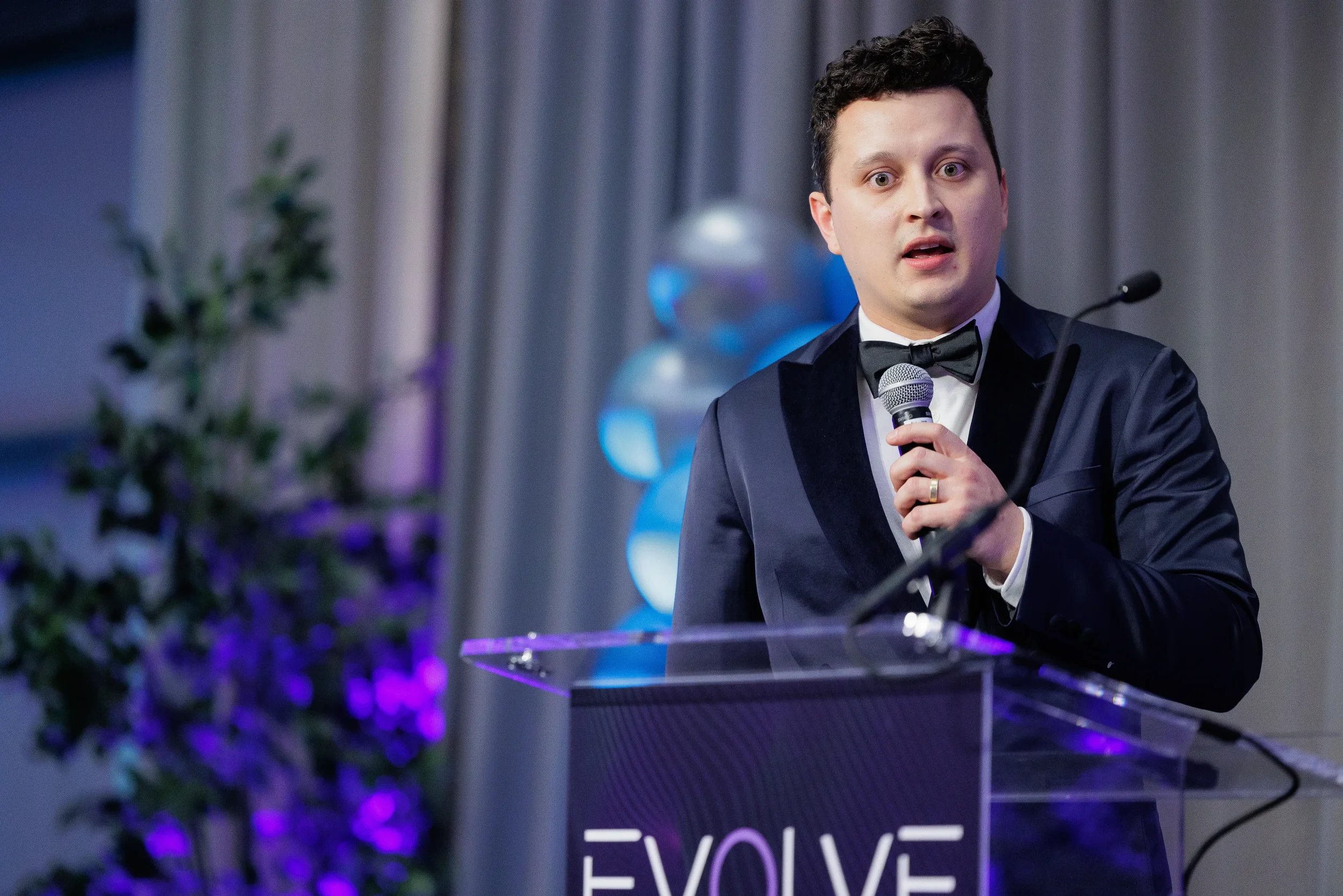 Tuxedo-clad emcee speaks into microphone at acrylic podium with balloon backdrop at Chicago corporate event