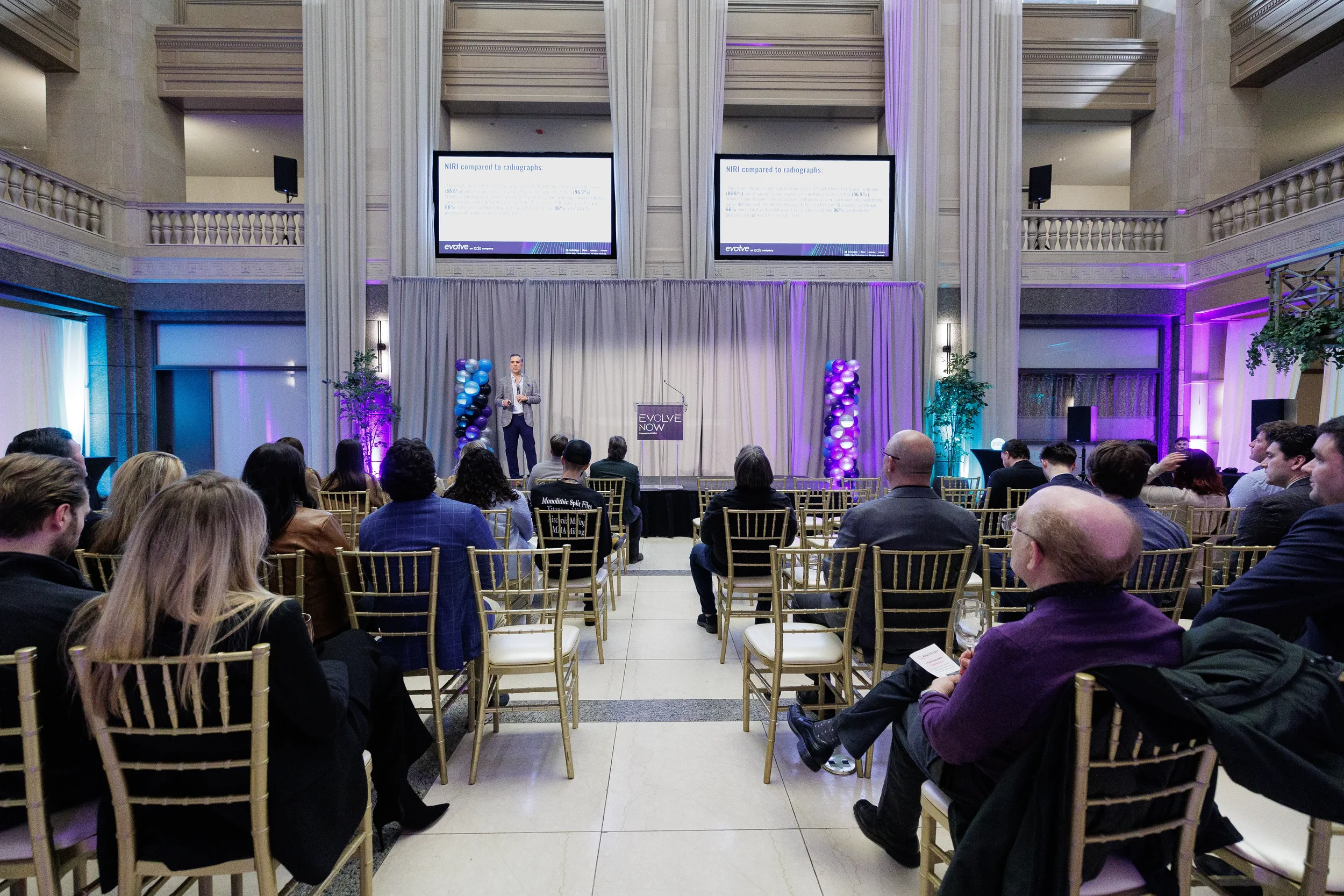 Wide view of seated audience watching speaker on stage with dual projection screens at Orlando industry conference