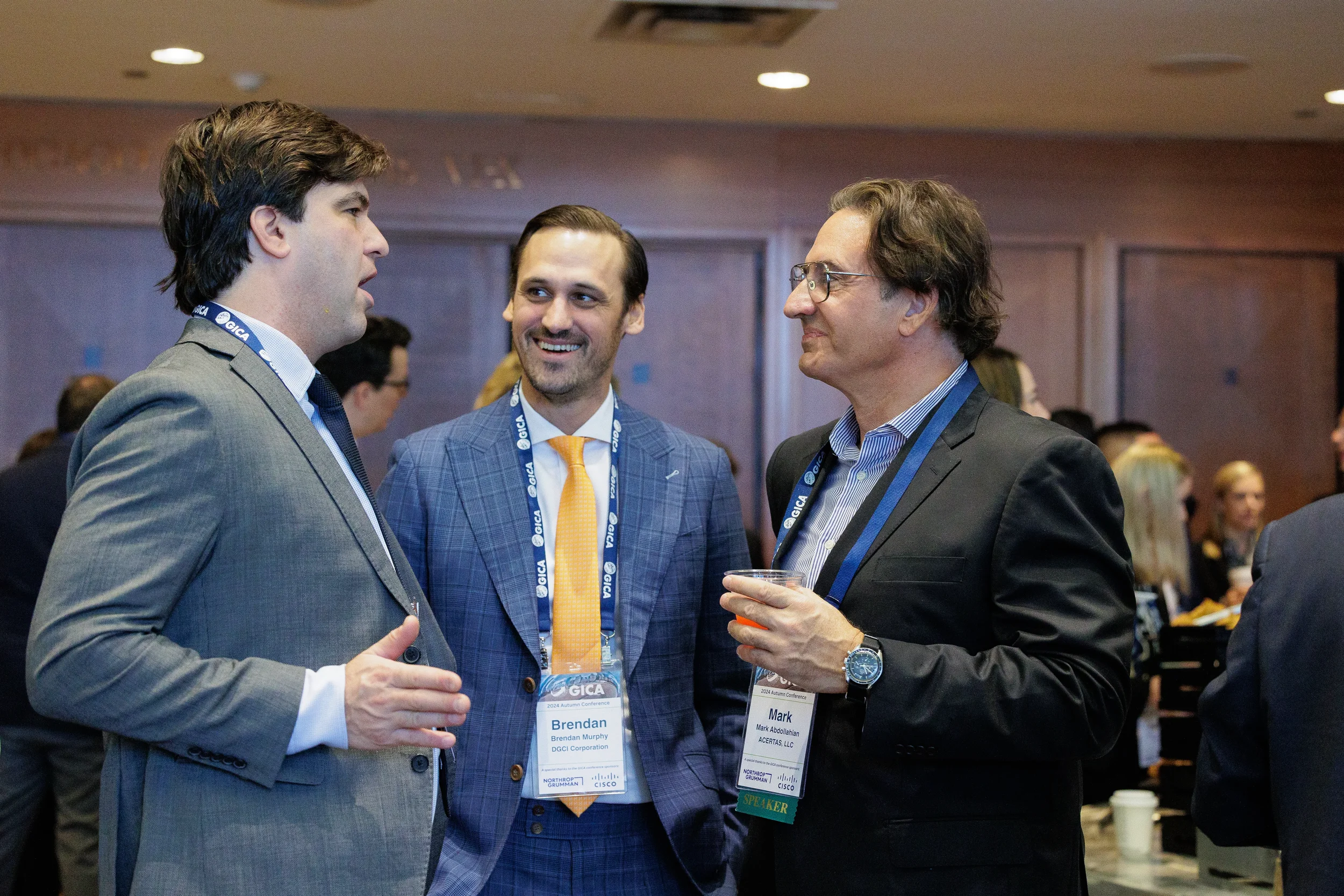 Three professionally dressed men wearing lanyards converse during a conference networking event in Chicago.