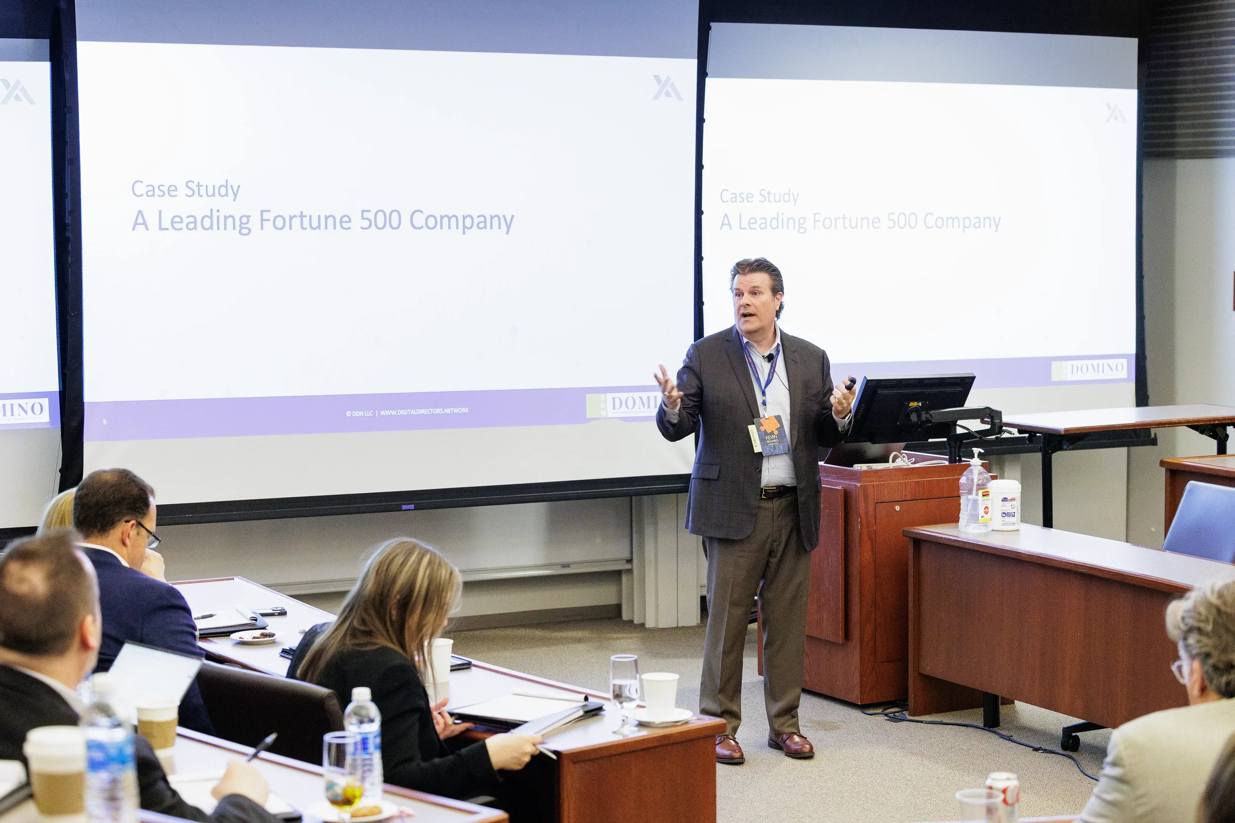 Male speaker gestures before tiered room audience with dual projection screens at Orlando corporate conference