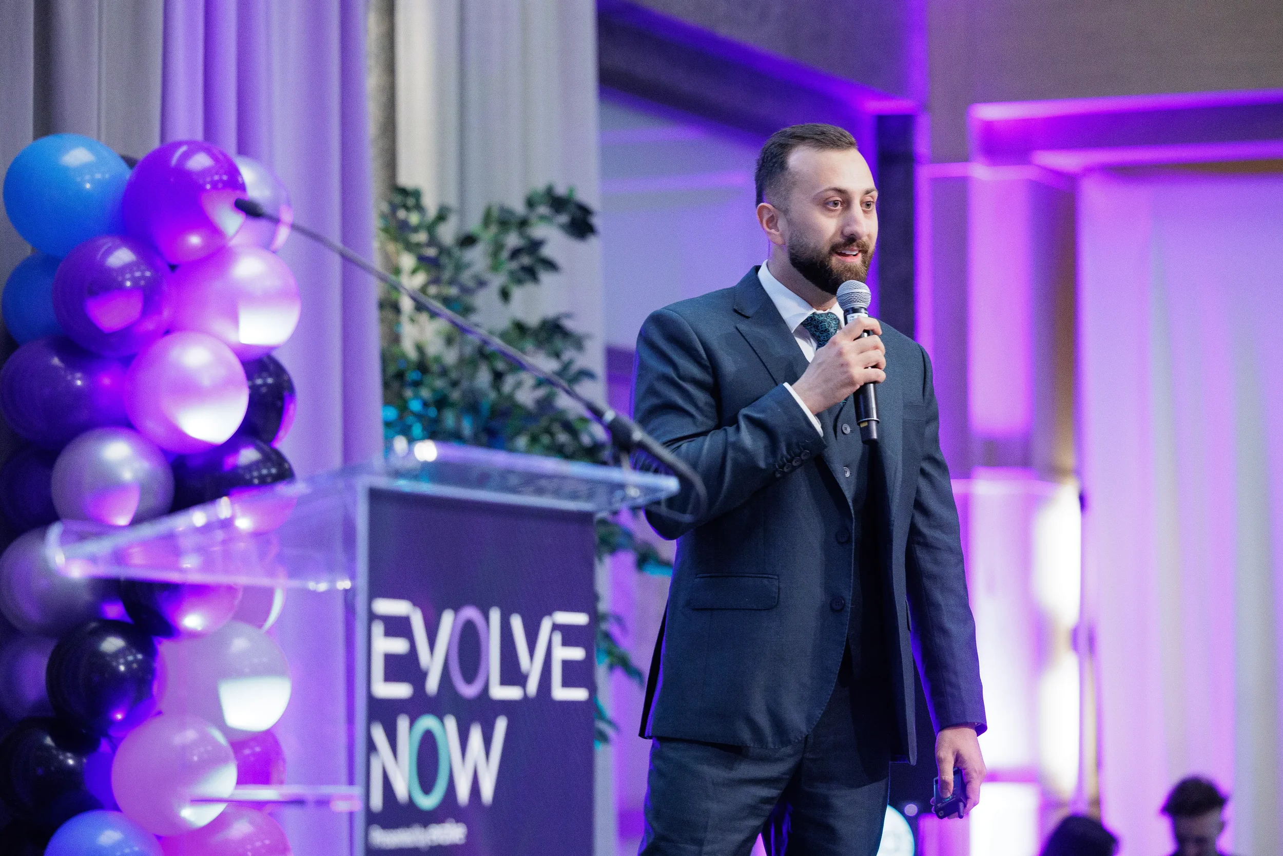 Three-piece suited speaker holds microphone beside podium with balloon column and purple uplighting at Chicago event