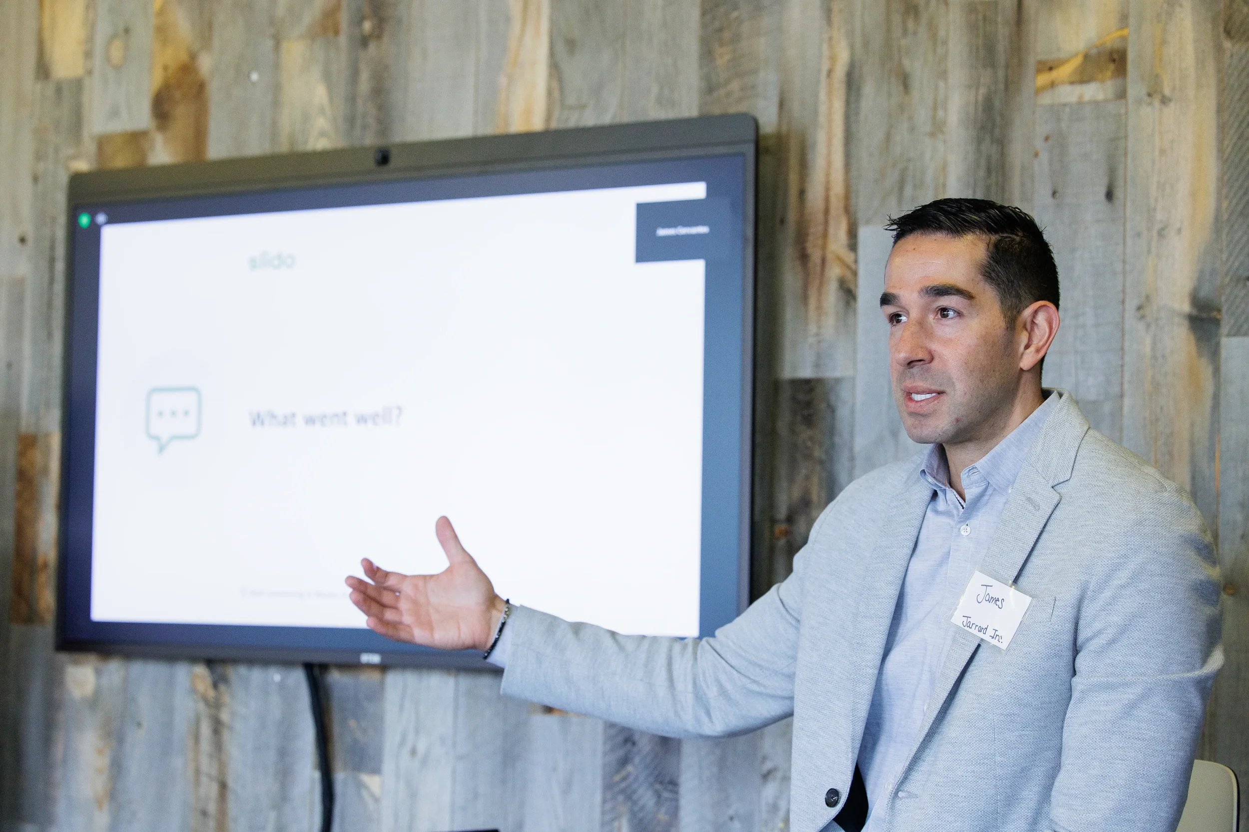 Male speaker gestures beside a display screen against a reclaimed wood backdrop at Orlando conference