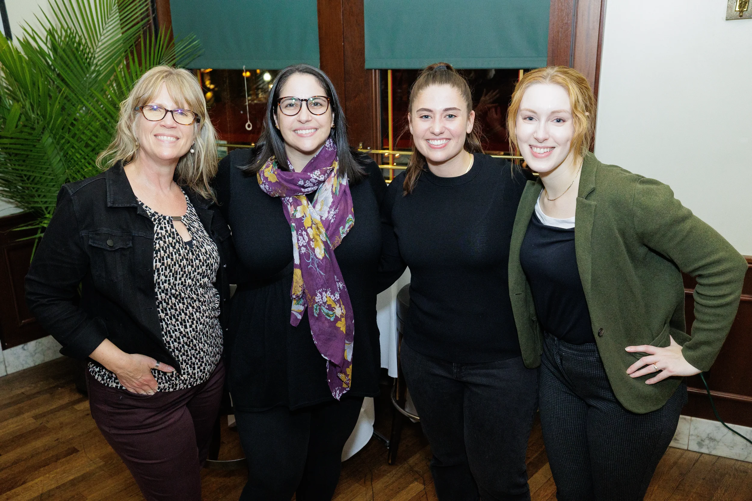 Four smiling women pose together indoors in front of dark wood paneling and a large green plant at a Chicago event.