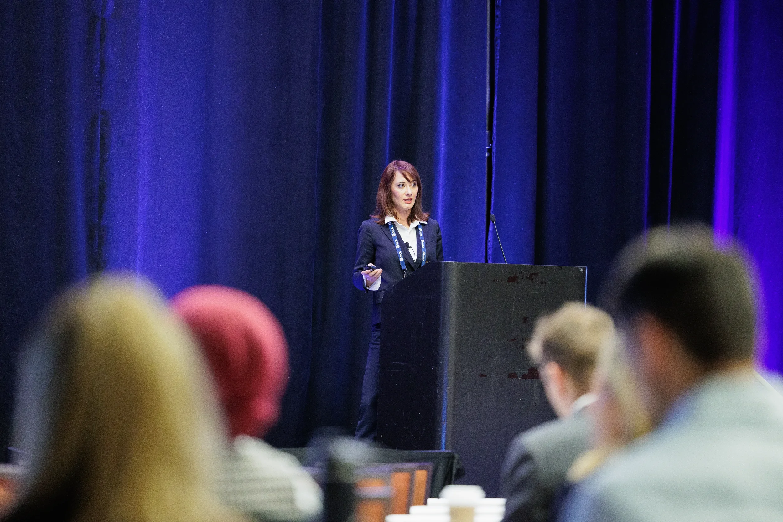 A business woman presents at a podium against a dark blue curtain, viewed over blurred audience members in Chicago