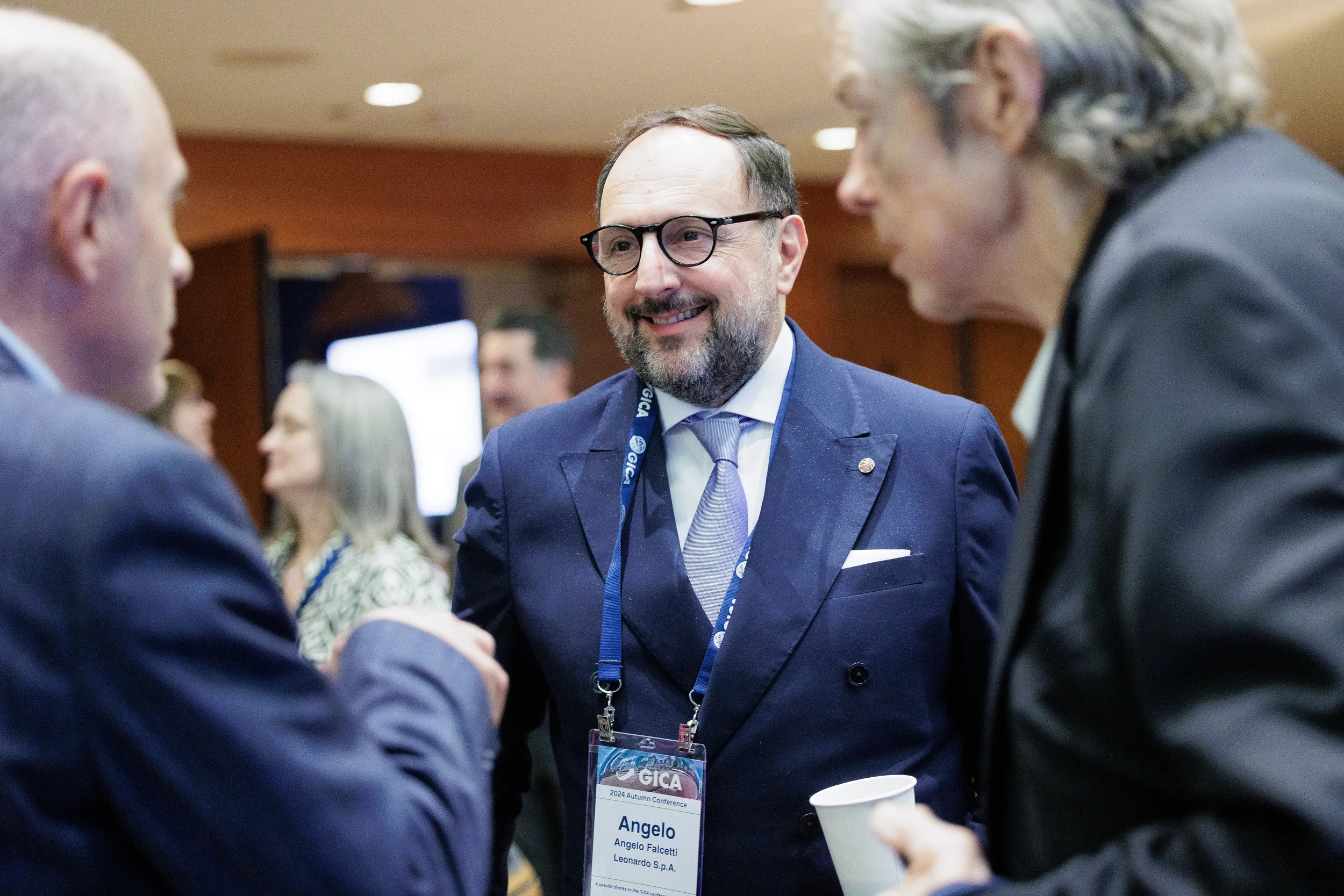 A smiling bearded man in glasses and navy suit talks with two other men at a conference in Chicago, wearing a GICA badge.
