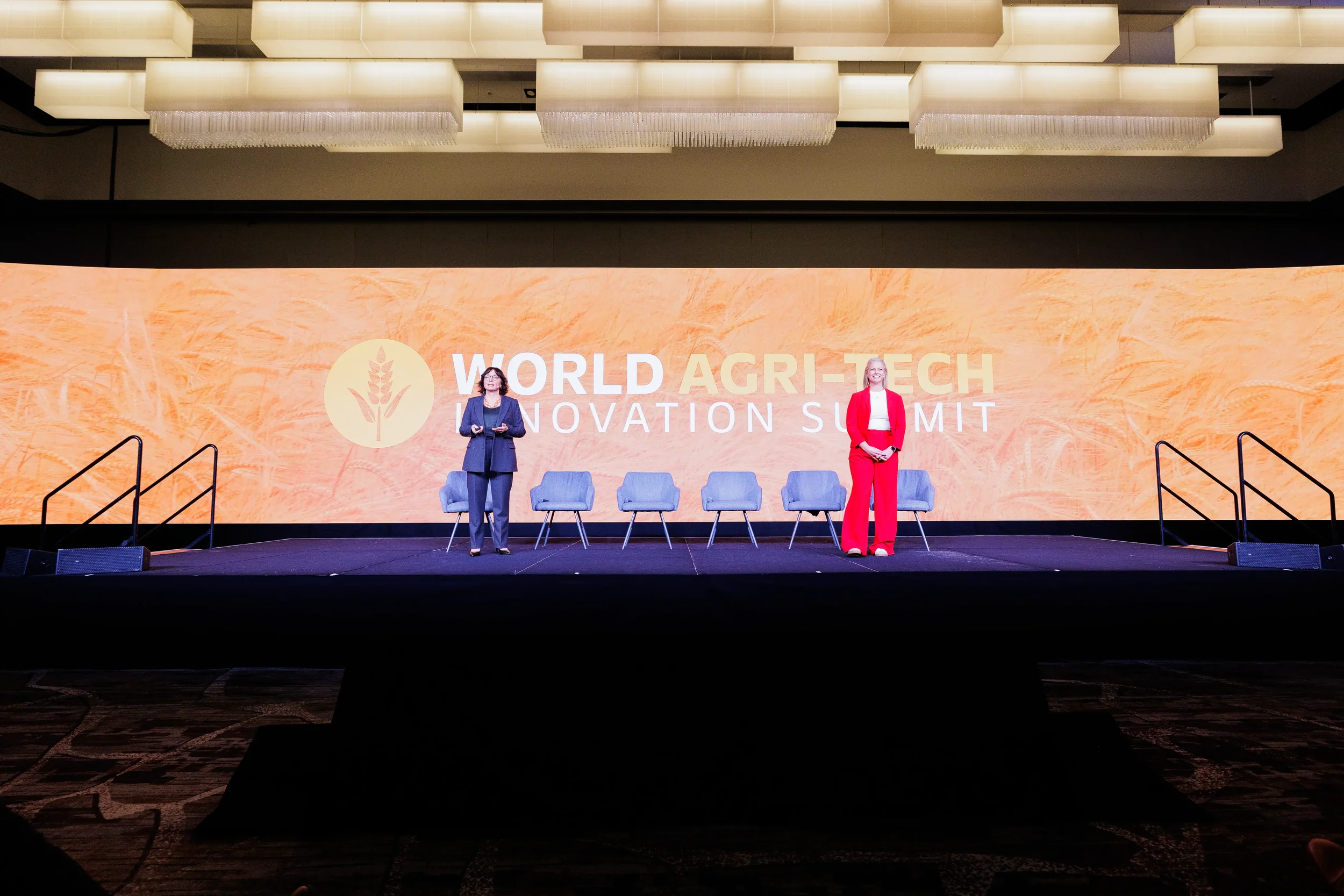 Two female speakers stand at opposite ends of wide stage before large branded LED backdrop at Orlando conference
