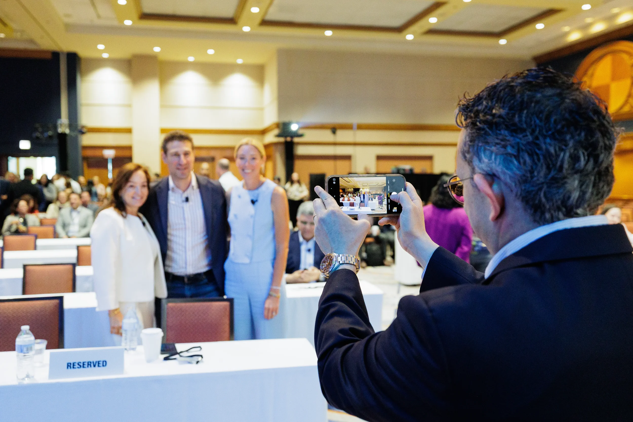 Attendee takes smartphone photo of three presenters posing in ballroom at Orlando corporate event