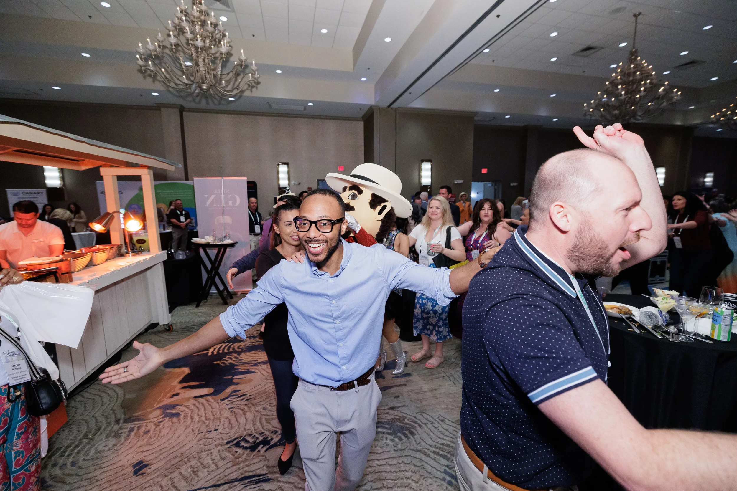 Attendees dance and celebrate with costumed mascot in chandelier ballroom at Orlando industry conference