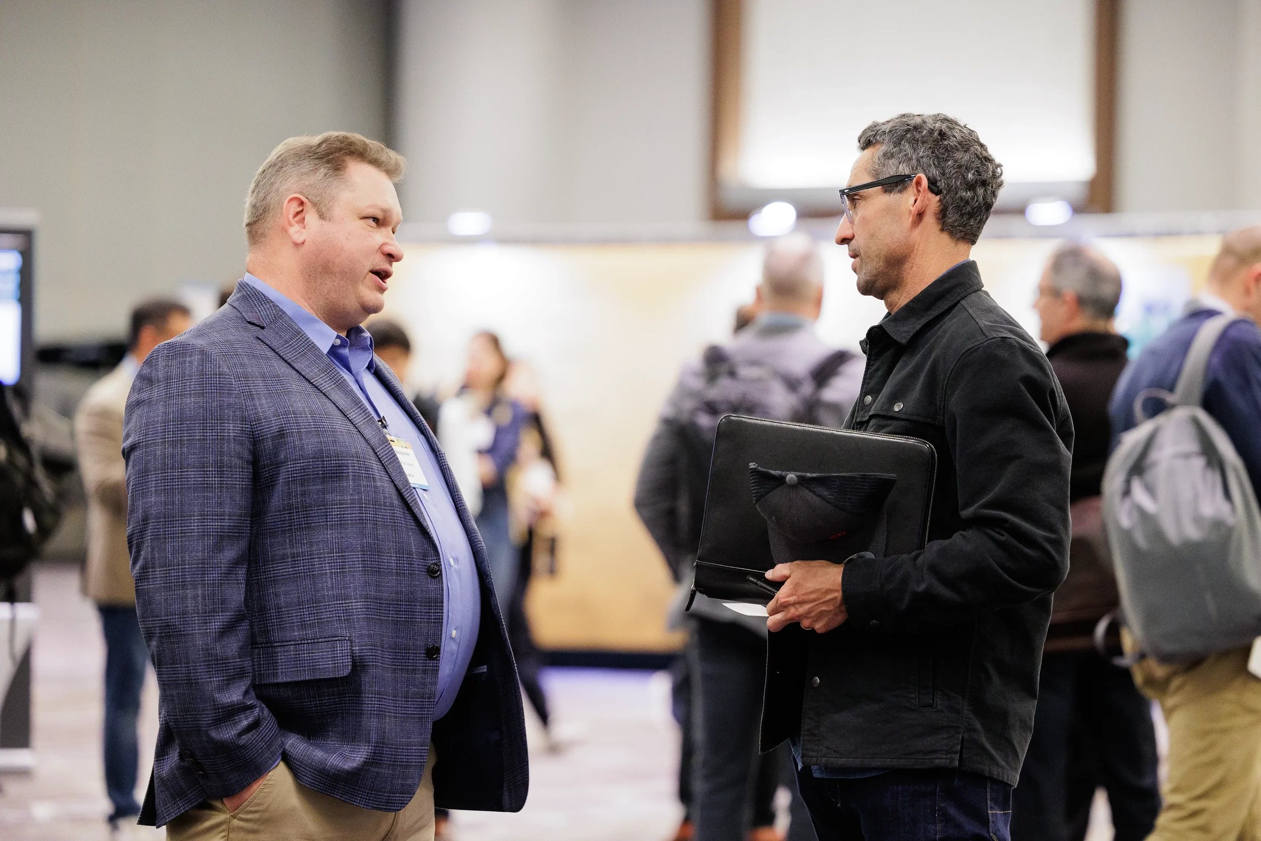 Two attendees in suits converse holding notebook during busy Chicago corporate event hallway