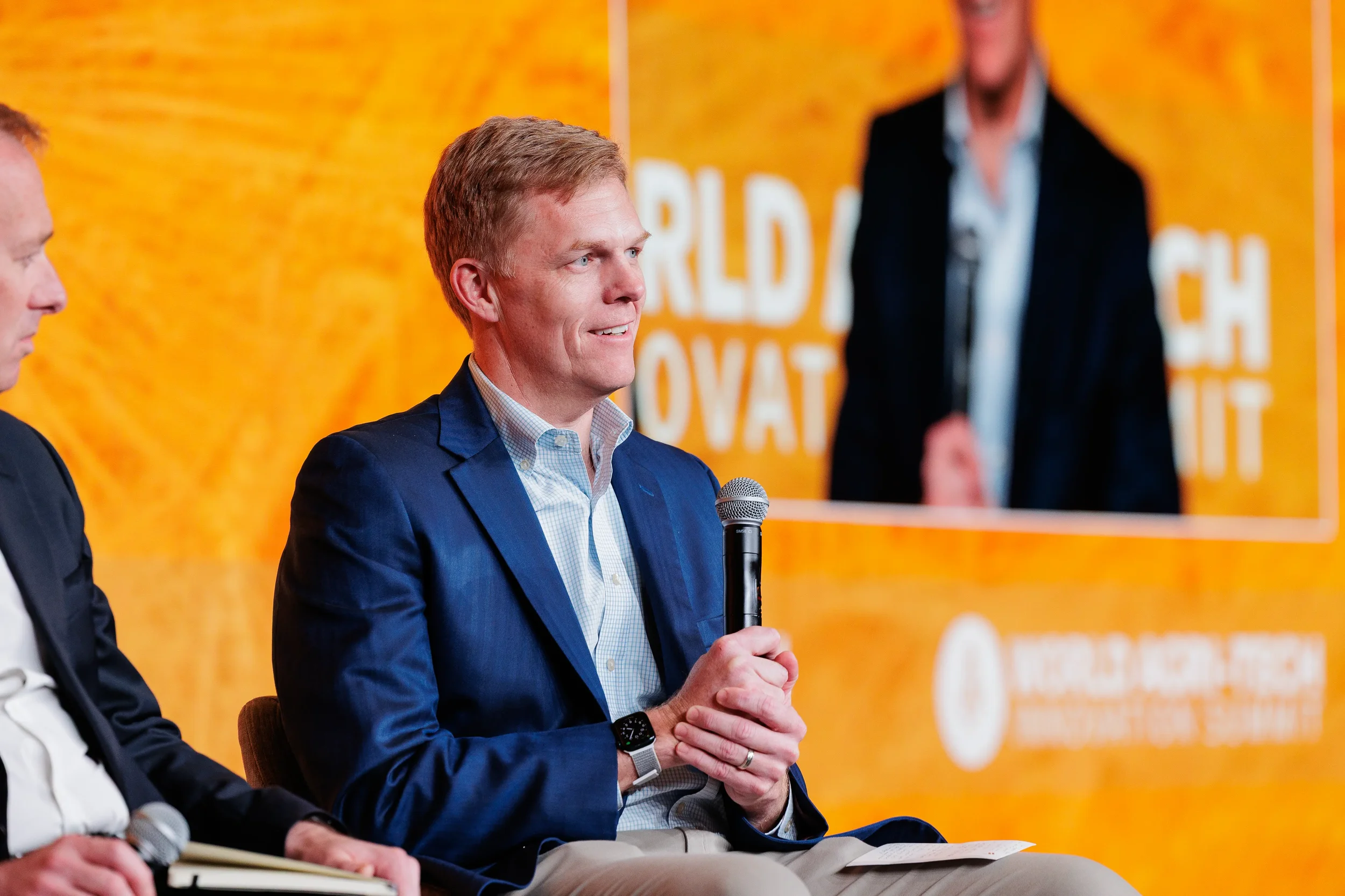 Male panelist in blue blazer holds microphone on stage before branded orange backdrop at Chicago conference