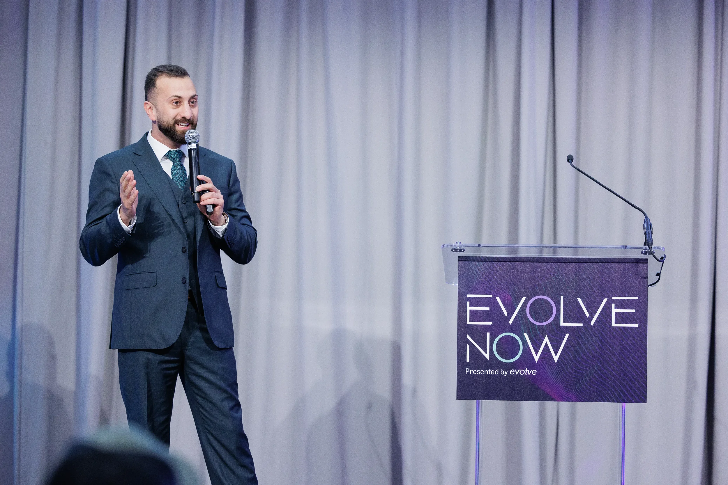 Smiling three-piece suited speaker presents beside branded podium on stage at Orlando corporate event