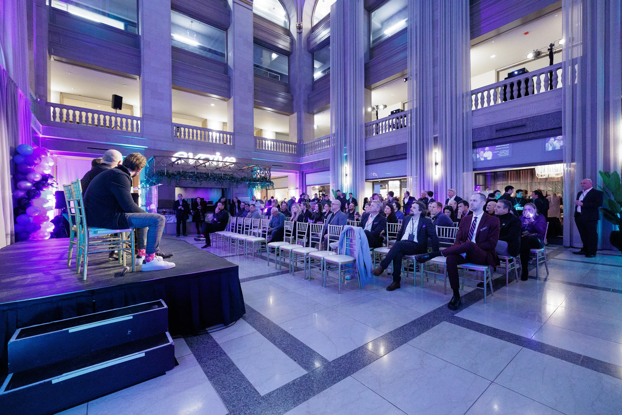 Two speakers seated on stage face engaged audience in grand atrium at Orlando corporate networking event