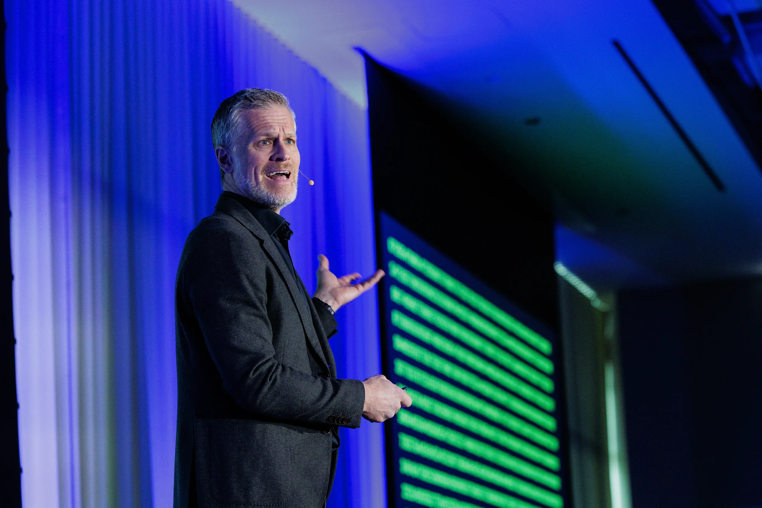 Speaker with headset microphone gestures toward green screen on stage at Chicago industry conference