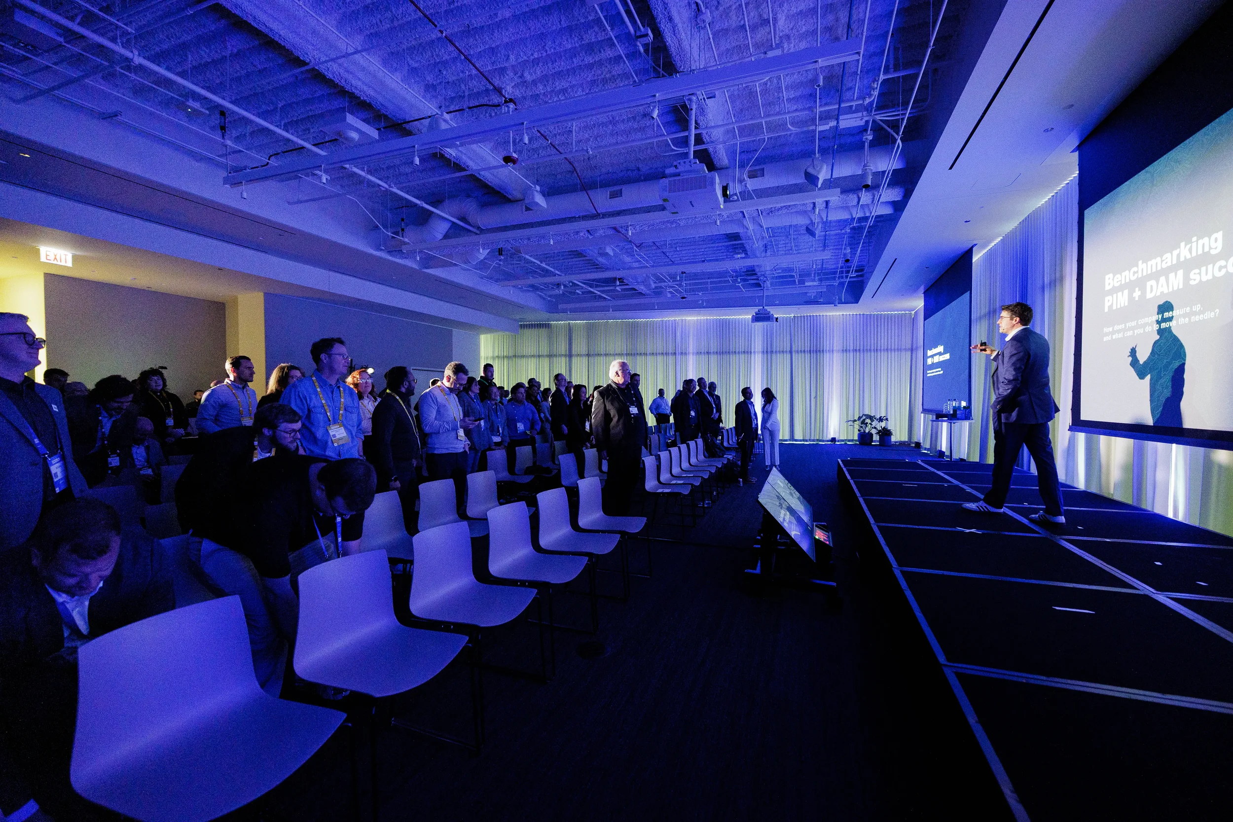 Speaker presents on stage as standing audience looks on in blue-lit room at Orlando industry conference