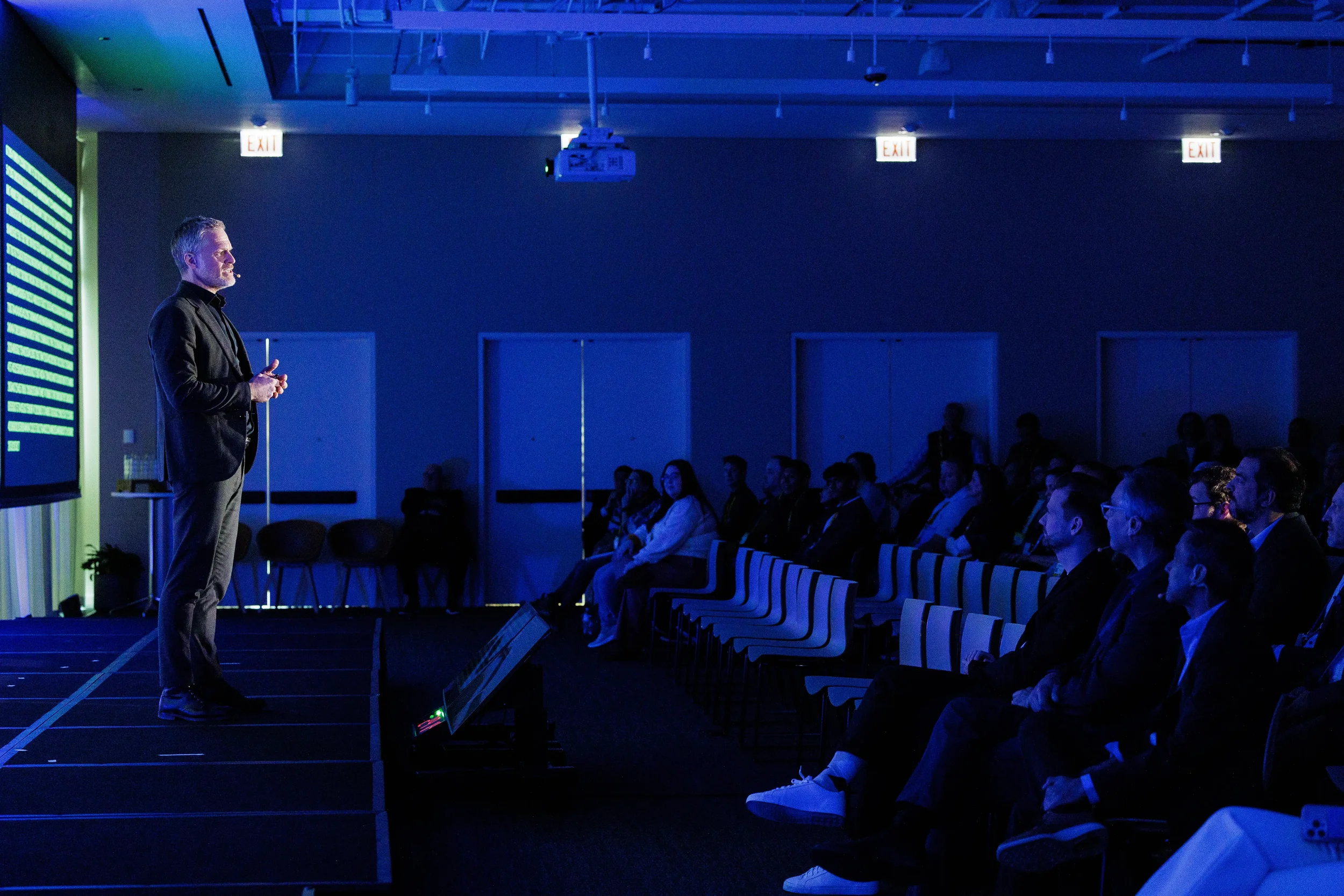 Speaker presents on stage to seated audience in blue-lit room during session at Orlando corporate conference