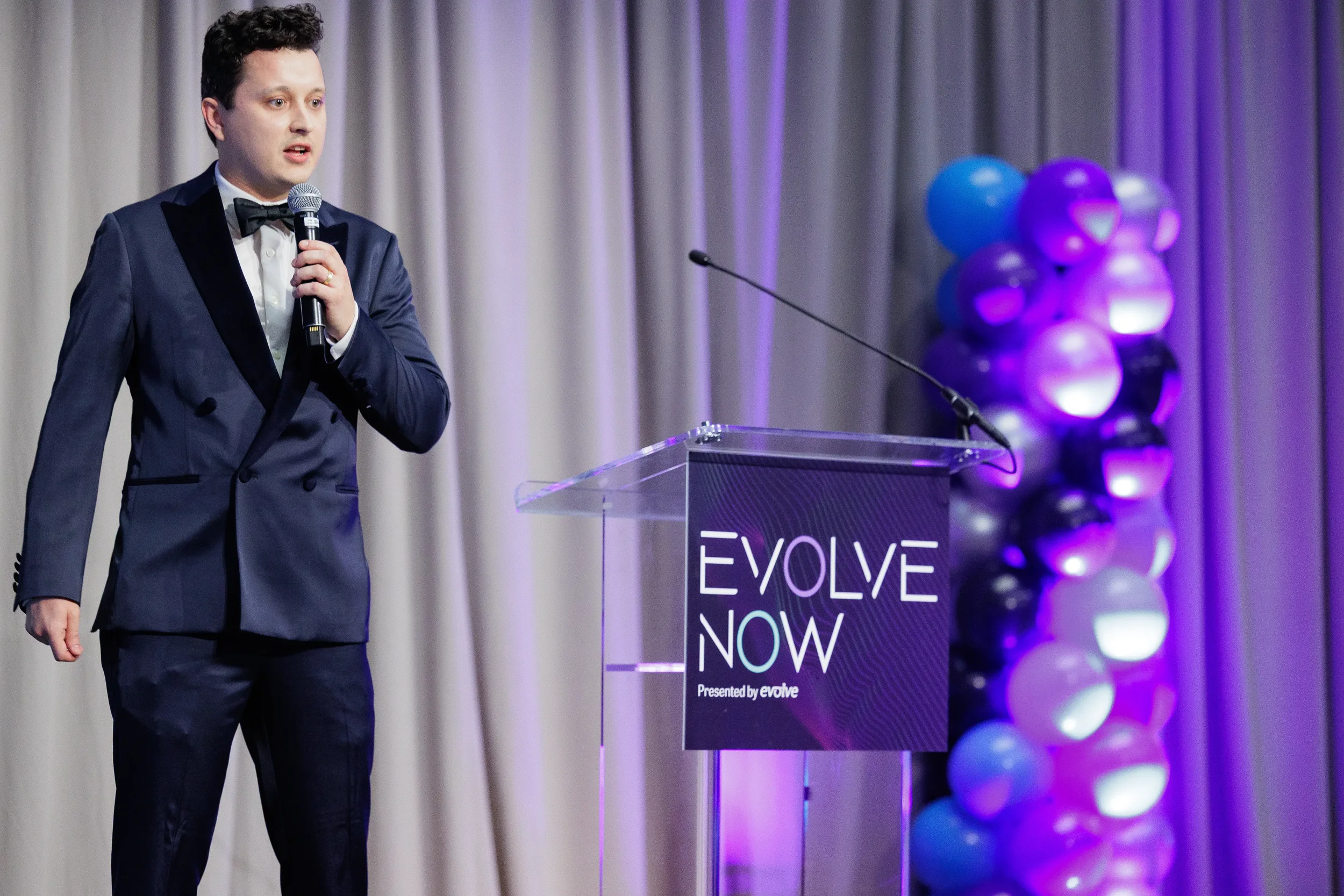 Tuxedo-clad speaker holds microphone beside acrylic podium with balloon column at Chicago corporate event