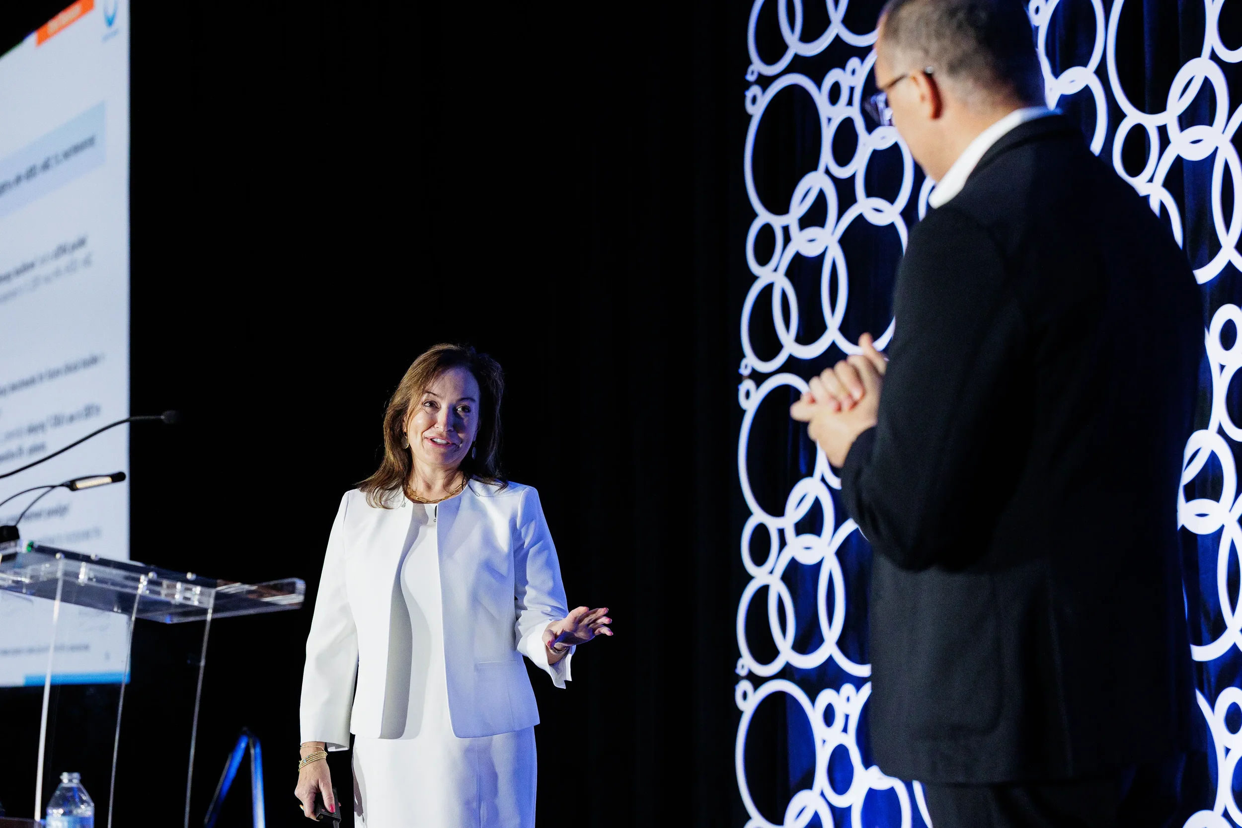 Presenter in white suit gestures on stage as colleague looks on at Chicago corporate meeting