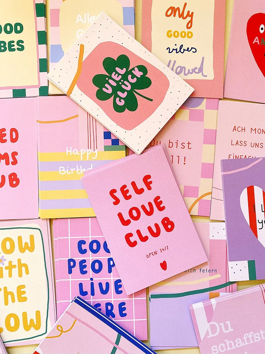 Eine Zusammenstellung von verschiedenen Postkarten und Notizzetteln mit bunten, freundlichen Botschaften wie "Self Love Club", "Joy & Luck", "Happy Birthday" und andere, in Pastellfarben und lebhaften Tönen.