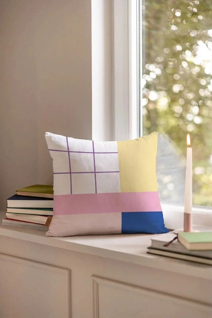 Bunte Kissen mit Pastellfarben auf einer weißen Fensterbank neben Büchern und einer Kerze, Blick nach draußen durch das Fenster, bei Tageslicht.