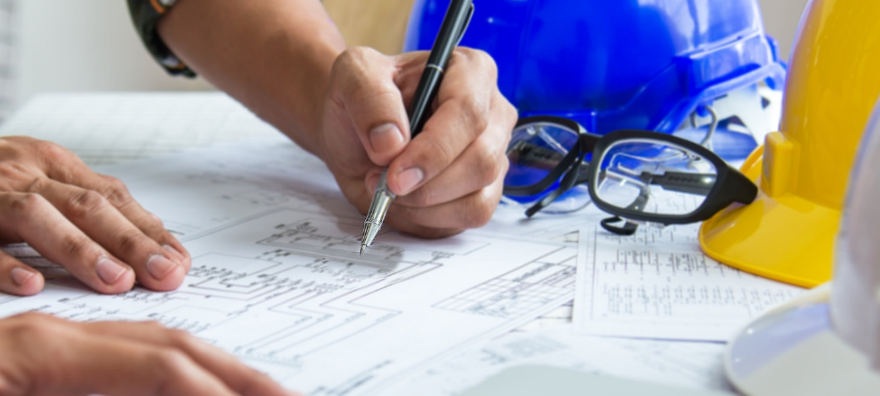 Close up of construction plans; man’s hands taking notes; hard hats on a table in the background.