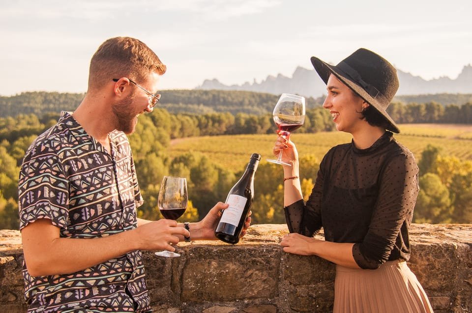A man and woman enjoying wine outdoors on a stone wall with scenic countryside in the background, the man holding a wine bottle and glass, the woman holding a glass of wine, both smiling.