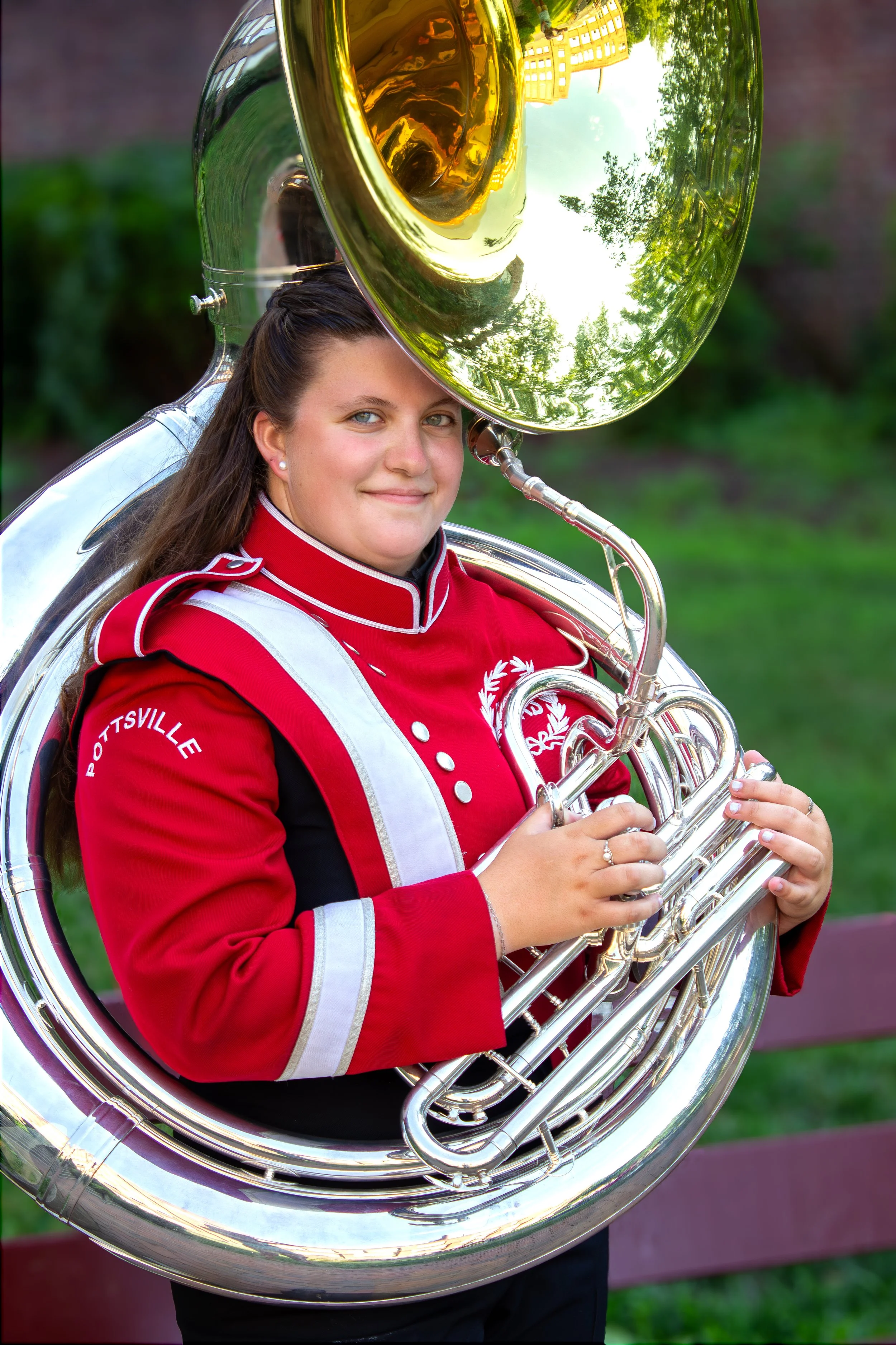 A high school senior girl in her red and white marching band uniform smiling while holding a sousaphone during an outdoor portrait session near Hershey, PA.