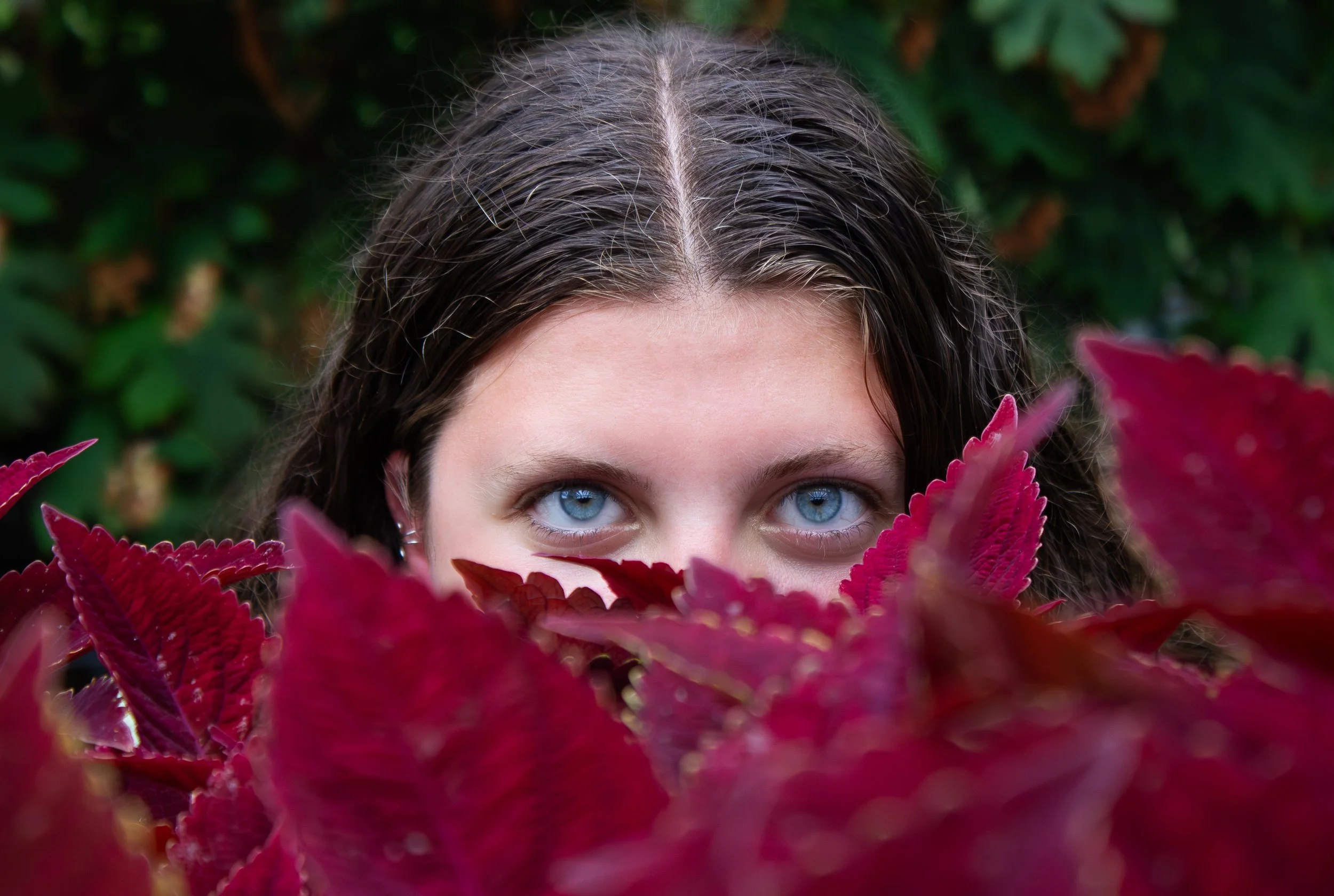 A woman peeking out from behind large red/burgundy leaves with striking blue eyes, set against green foliage.