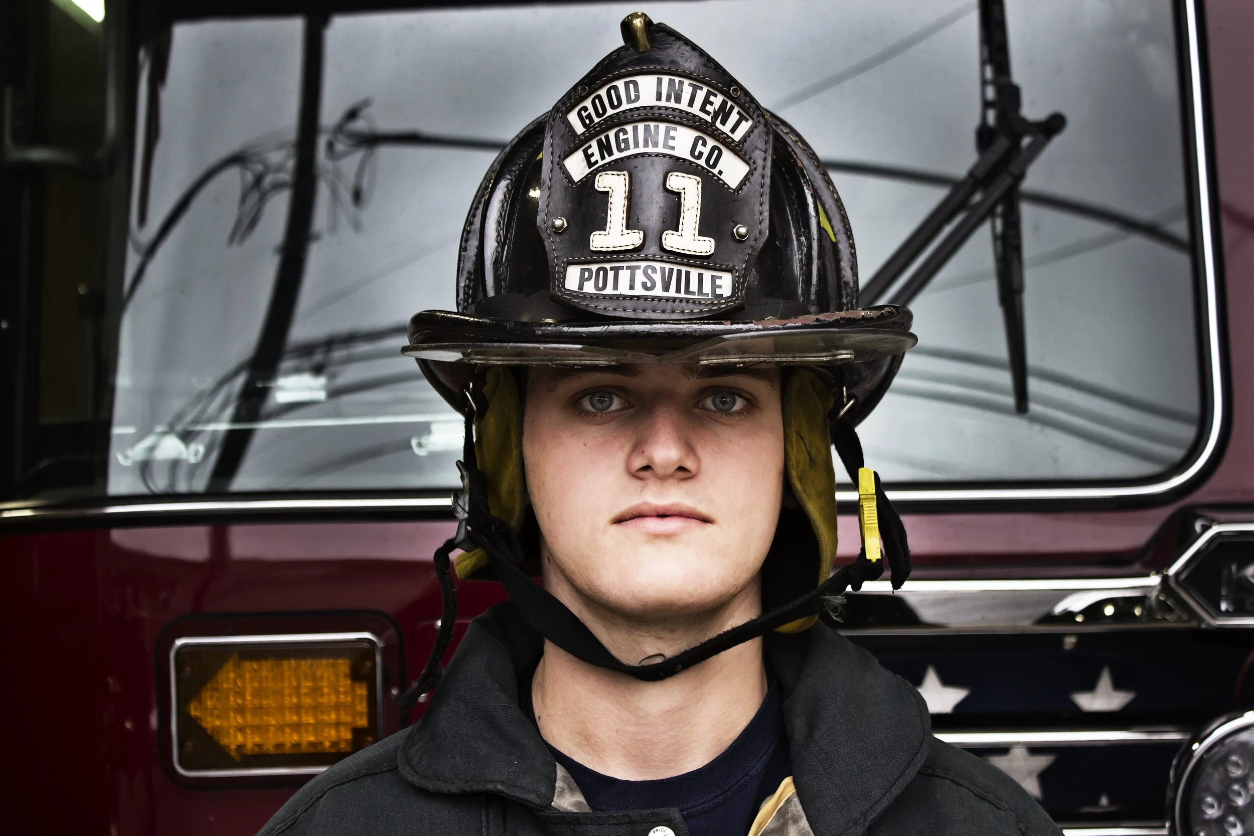 A close-up, authentic senior portrait of a young man wearing a Good Intent Engine Co. firefighter helmet, showcasing a natural expression rather than a forced pose.