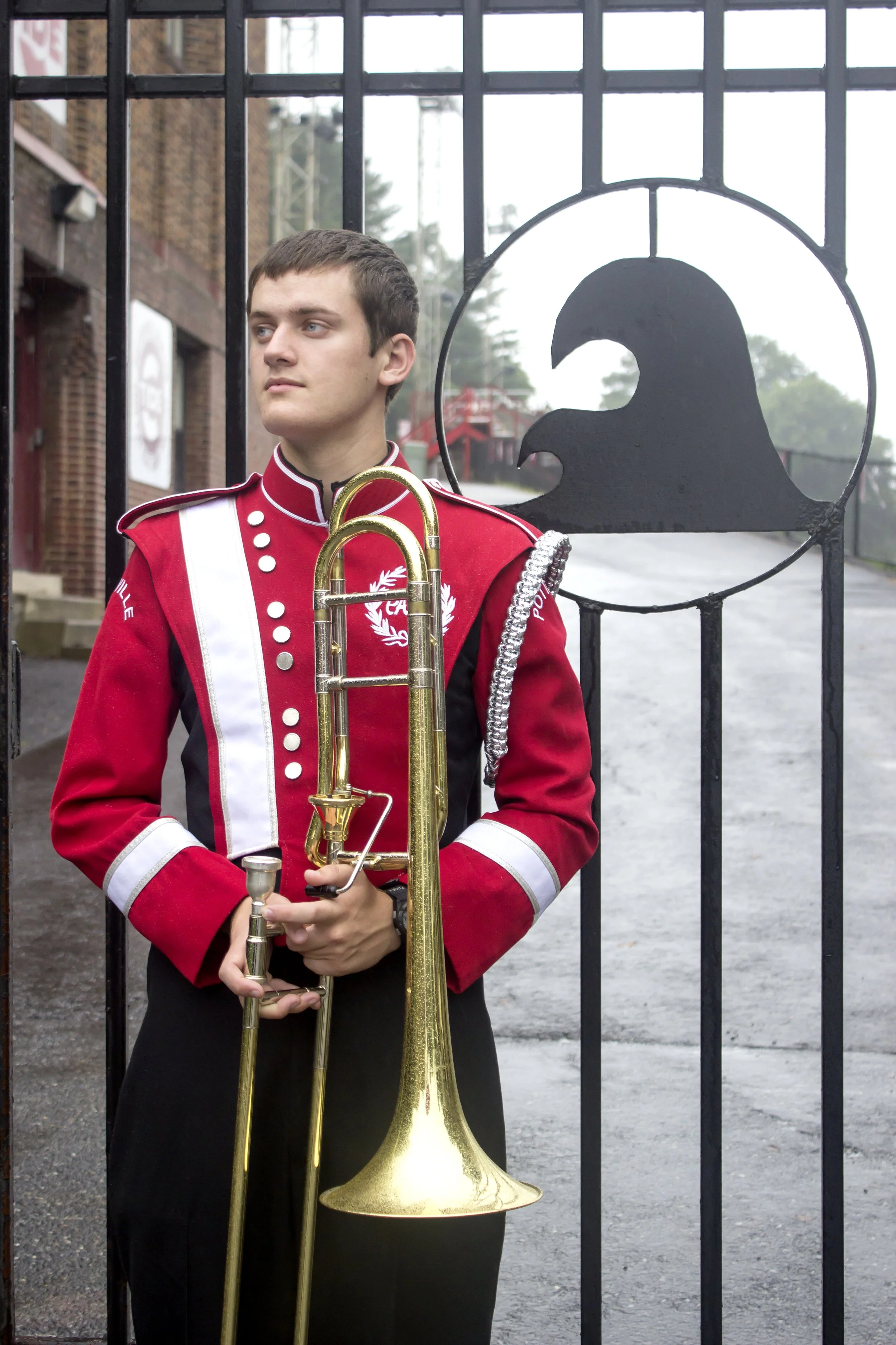 A male high school senior in a red and white marching band uniform holding a trombone while standing in front of a school gate, capturing his personal interests through an authentic portrait session.