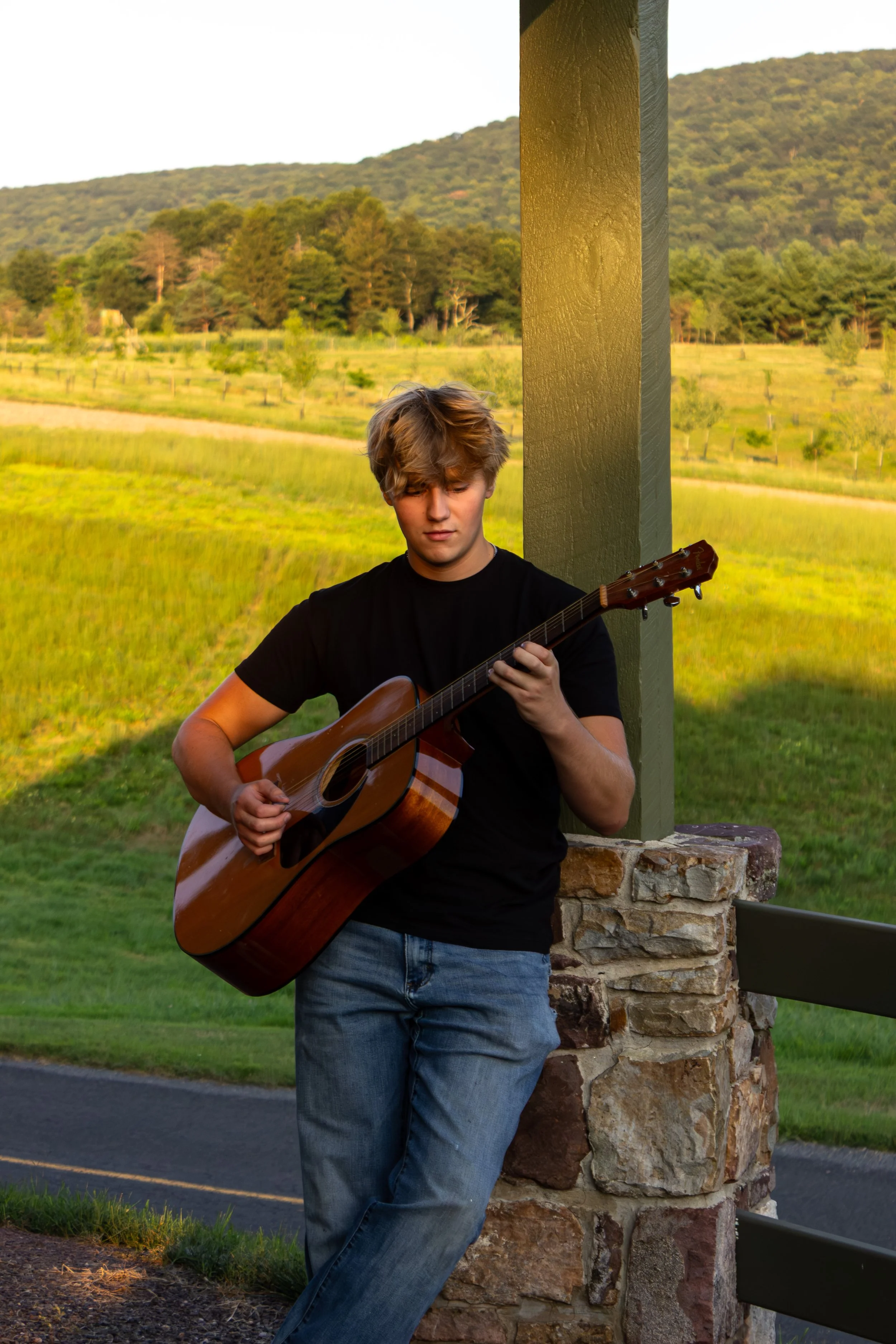 A high school senior boy sitting outdoors in a field, authentically captured playing an acoustic guitar during a story-driven portrait session in New Cumberland, PA.
