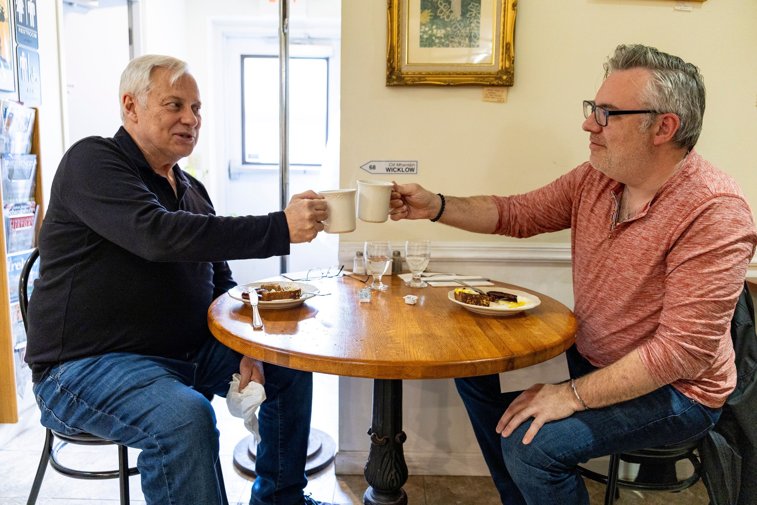 Two men smiling and toasting coffee mugs at a wooden table inside Oxford Hall Celtic Shop and Cafe.