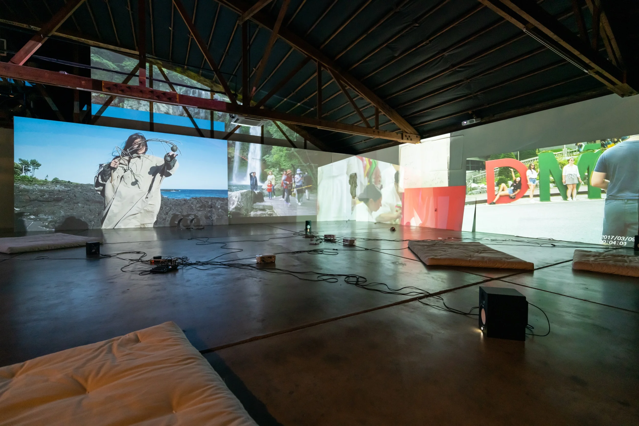 Dylan Mira, as if i was a thing i could do in the dark , installation view 3 channel HD video installation, sound, bells, fake Issey Miyake trenchcoat made in Korea, hat, plexiglas, paper, futons 12:00, looped Disjecta Contemporary Arts Center, curated by Suzy Halajian, 2019
