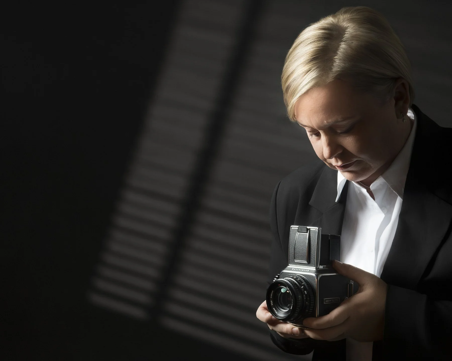 A woman in a black suit and white shirt holding a vintage camera, looking down at it, with a dark, blurred background.