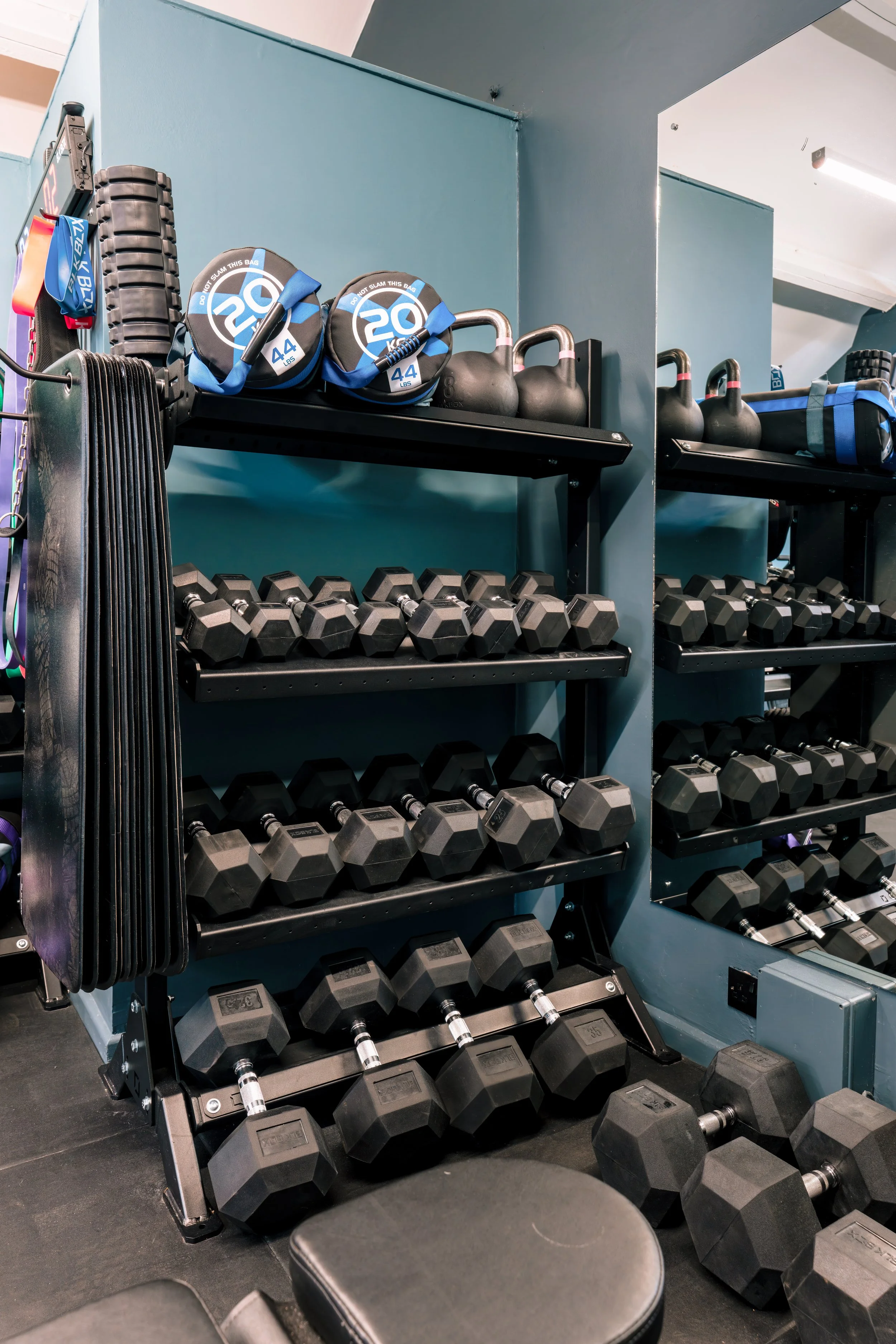 Night gym with black dumbbells on racks, kettlebells, weight bags, and additional weights on black shelves against teal walls.